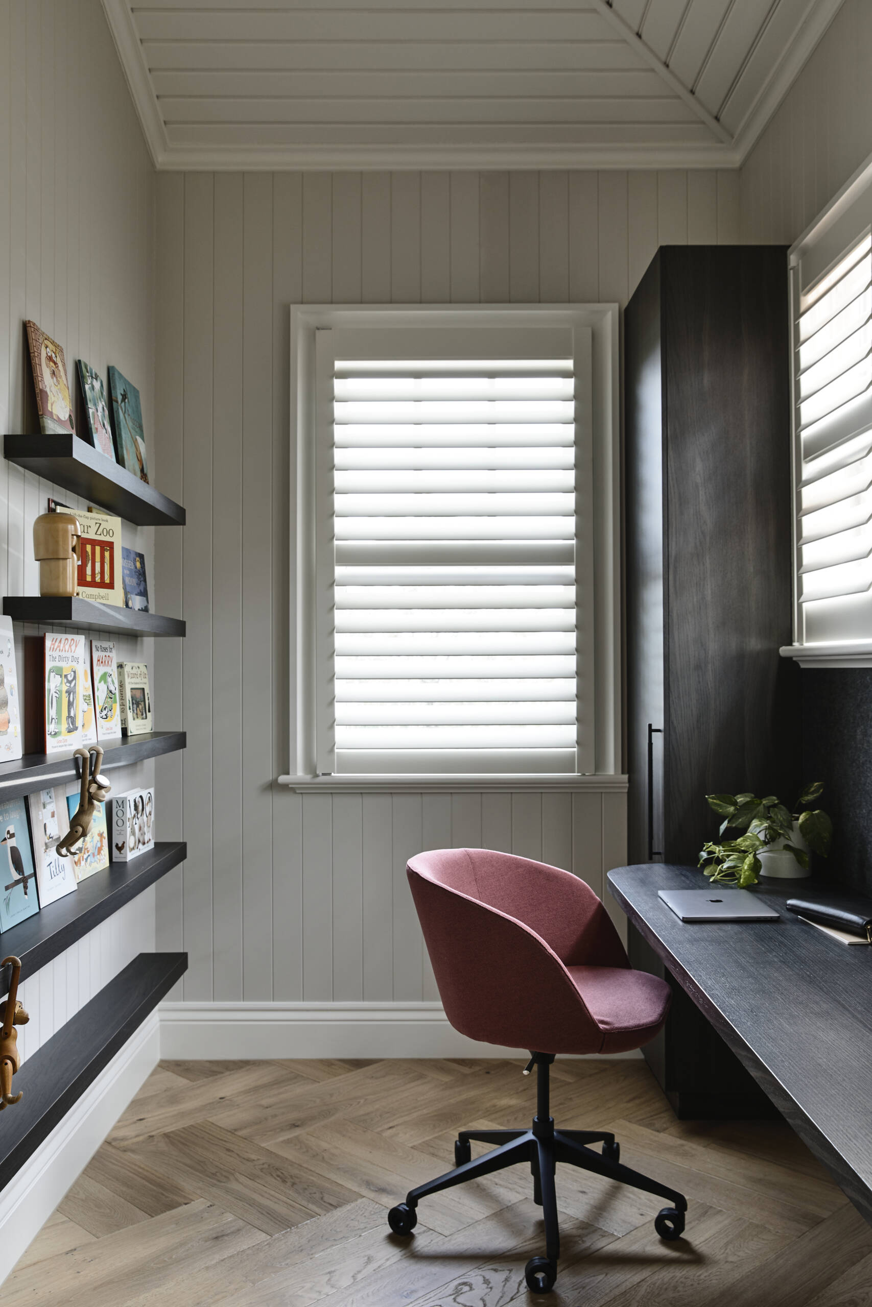A compact home office with soft gray beadboard walls, white shutters, and warm timber floors. Floating shelves and a dark wood desk create an organized, personal workspace.