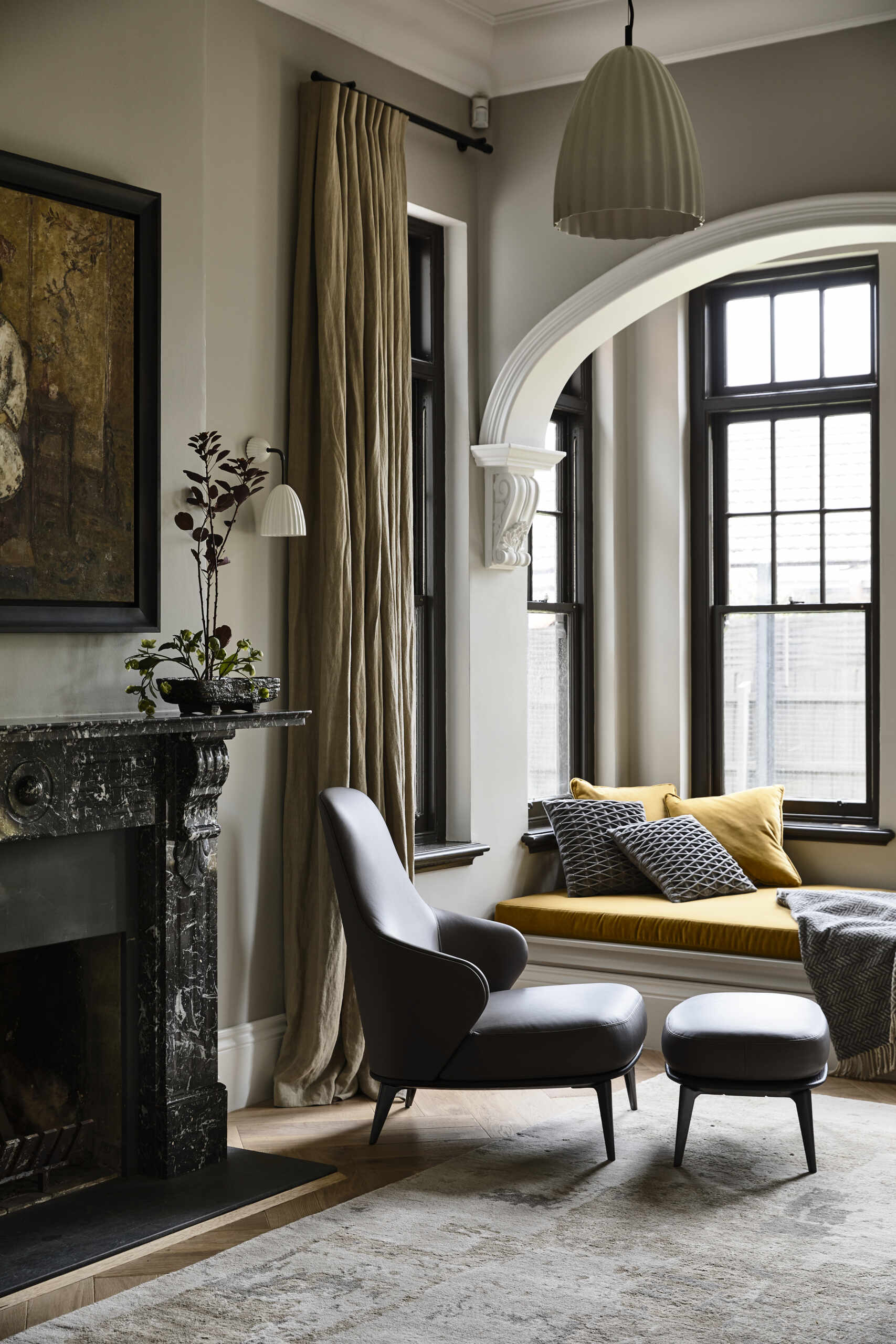 A calm living room anchored by a dark stone fireplace and softened with layered seating. An arched opening frames a built-in window seat that draws light into the space.