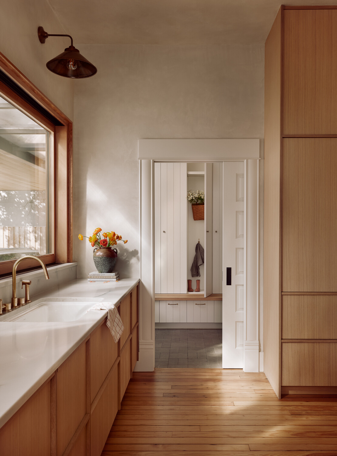 Visible through the doorway off the kitchen, the mudroom features built-in white paneled cabinets, paired with a simple wood bench that creates an easy spot to sit and slip off shoes. Low drawers beneath the bench keep everyday storage tucked away, while hooks and a small hanging basket add just enough function without visual clutter.