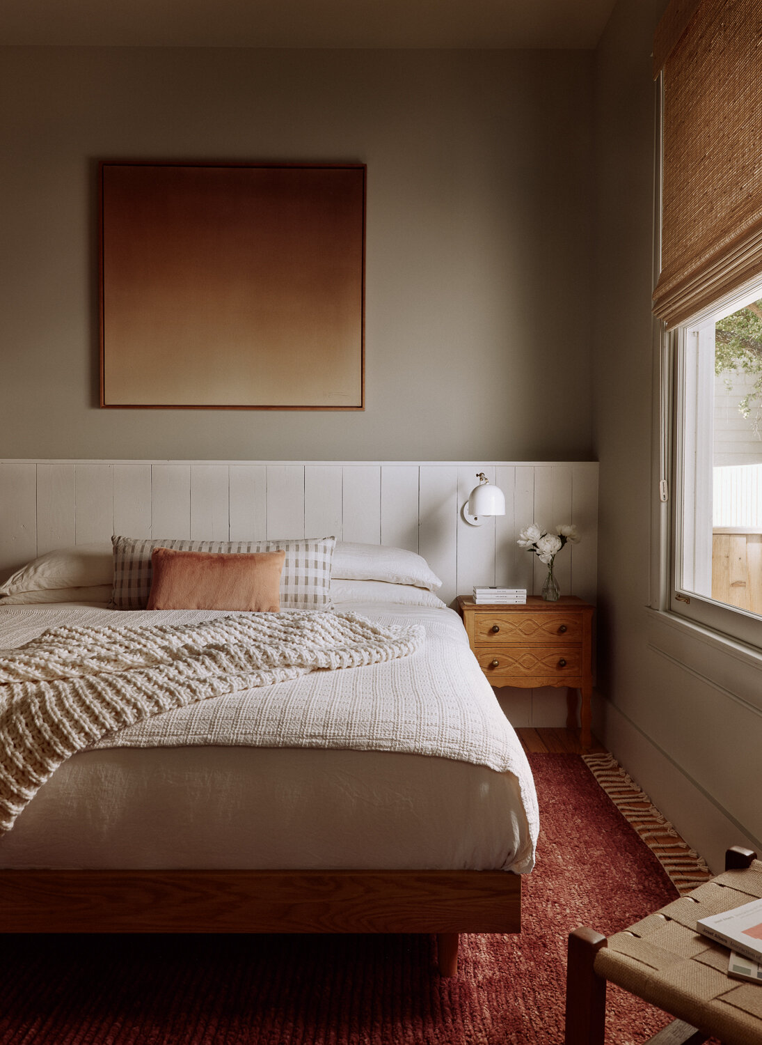 The primary bedroom leans into a sense of quiet and ease. Soft neutrals and warm, earthy tones set a calming foundation, while textured details like a chunky knit throw, gingham pillows, and a blush accent cushion add warmth without clutter. Painted wall paneling introduces subtle texture and a vintage nightstand with simple white sconce above keep the room grounded and functional.