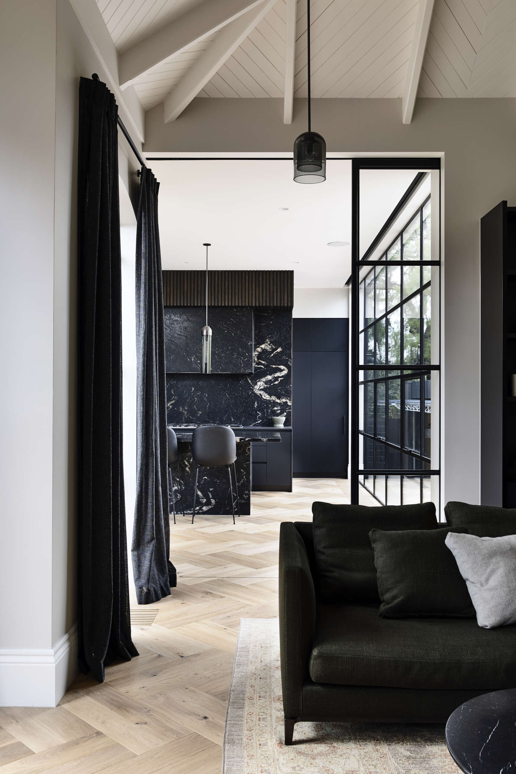 A sleek kitchen with matte black cabinetry, ribbed timber details, and dramatic black stone surfaces. Oversized grid windows and herringbone wood floors bring warmth and garden views inside.