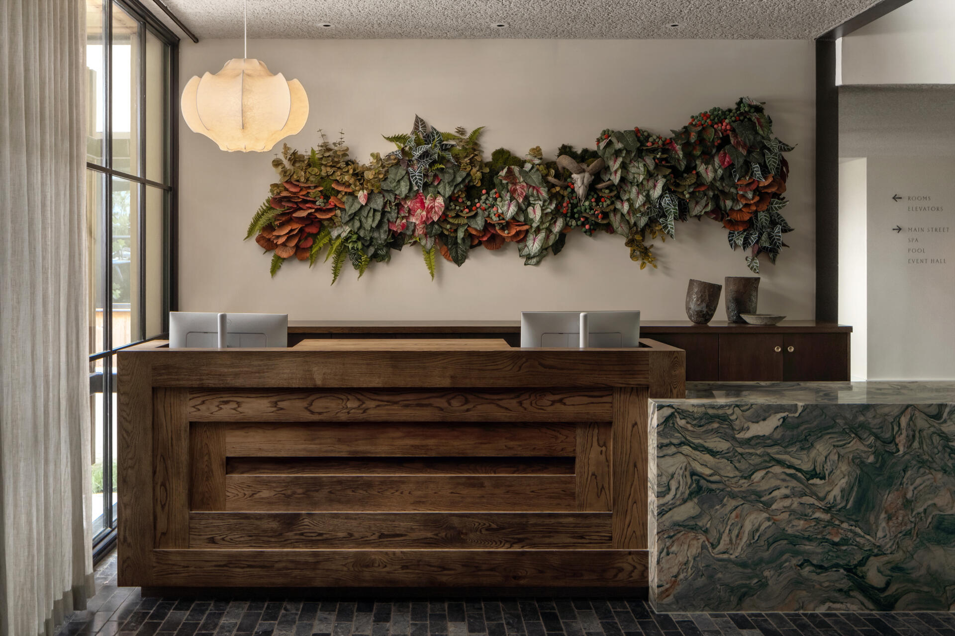 This hotel lobby unfolds as a series of connected spaces that balance warmth with quiet character. A sculptural wood reception desk sits against green stone and soft pendant light, while a plant accent adds a subtle sense of life.