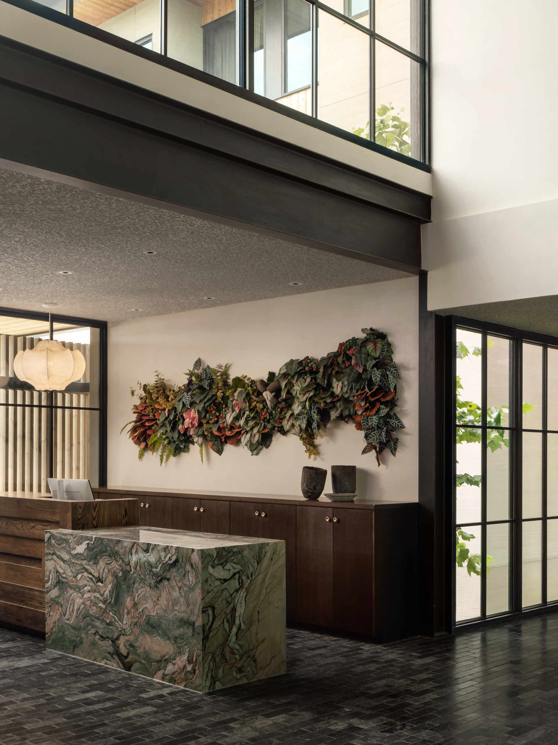 This hotel lobby unfolds as a series of connected spaces that balance warmth with quiet character. A sculptural wood reception desk sits against green stone and soft pendant light, while a plant accent adds a subtle sense of life.