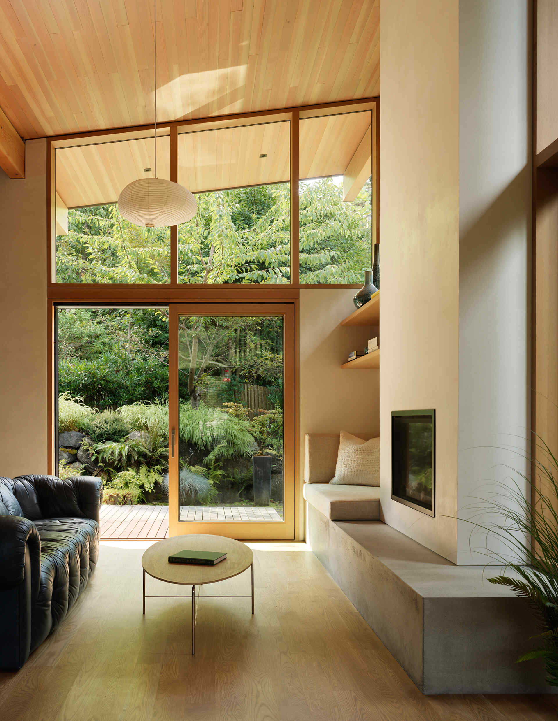 The living area is anchored by a wood-burning fireplace beneath a soft loft, offering warmth and focus. Expansive windows draw in light and frame views of moss-covered rockery, ferns, and native trees. A cozy seating nook lets residents enjoy quiet moments while staying connected to the outdoors.