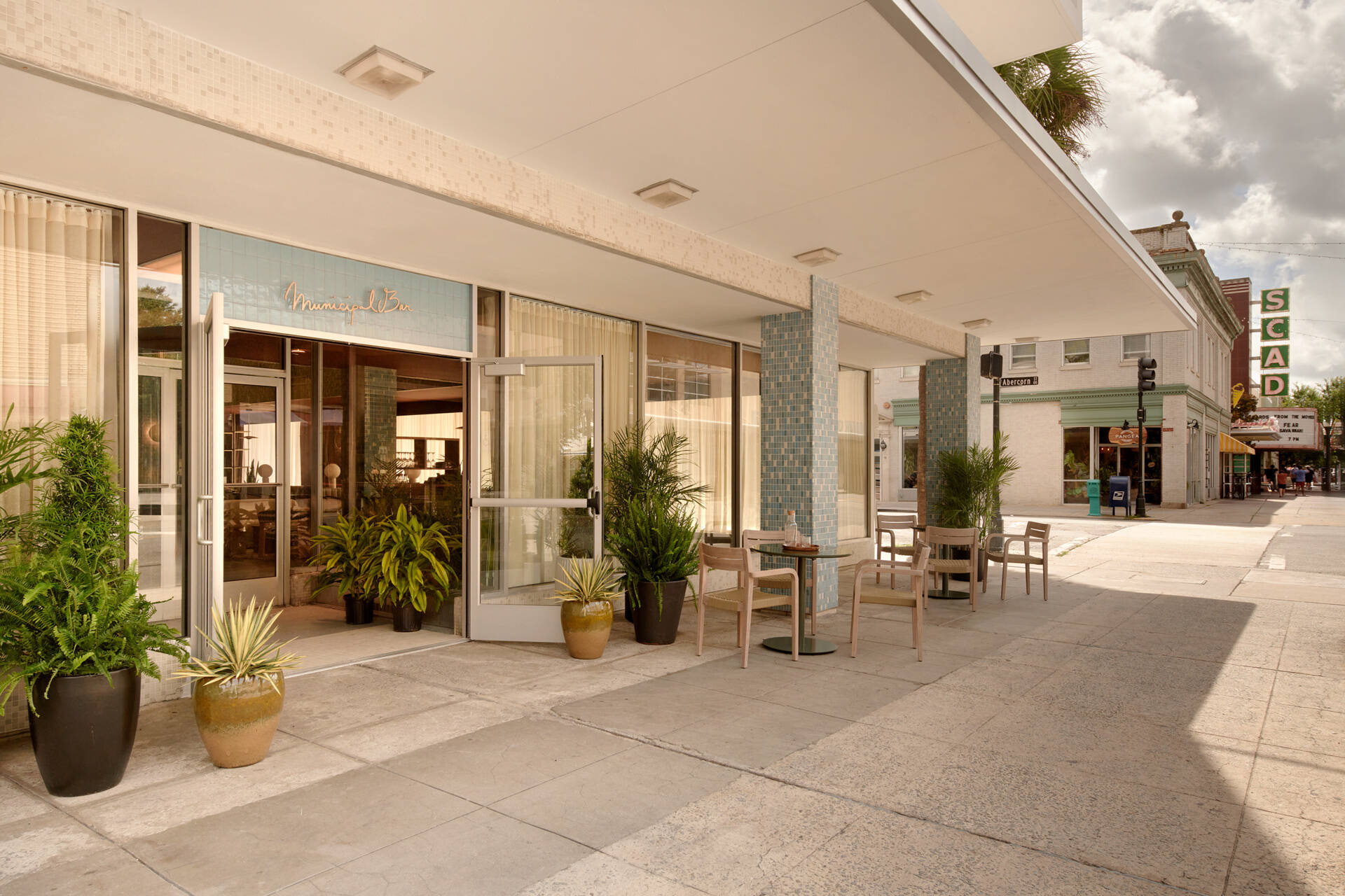 A softened street-facing hotel entrance uses lighting, seating, and planters to welcome guests into Municipal Grand from busy downtown Savannah.