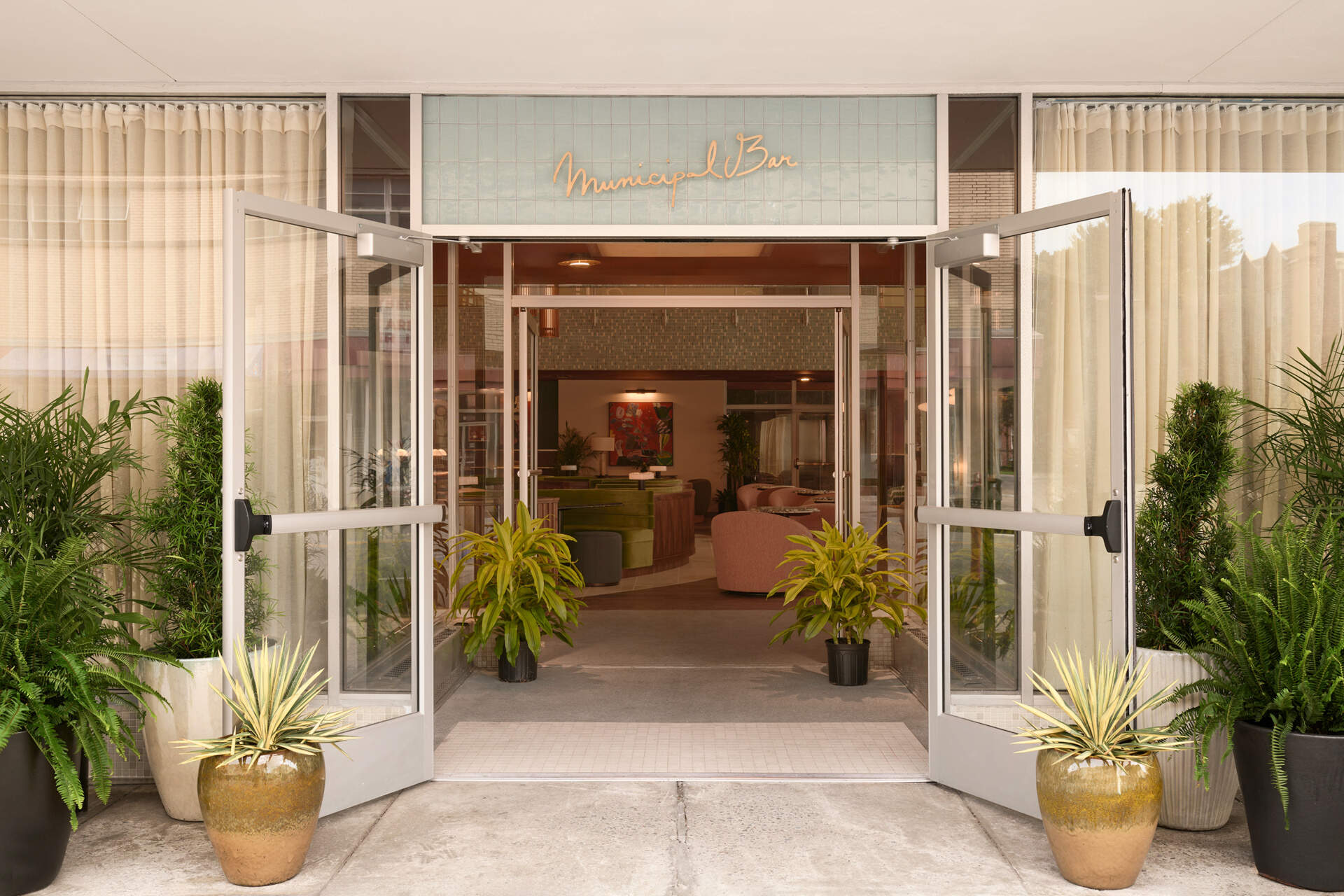 A softened street-facing hotel entrance uses lighting, seating, and planters to welcome guests into Municipal Grand from busy downtown Savannah.