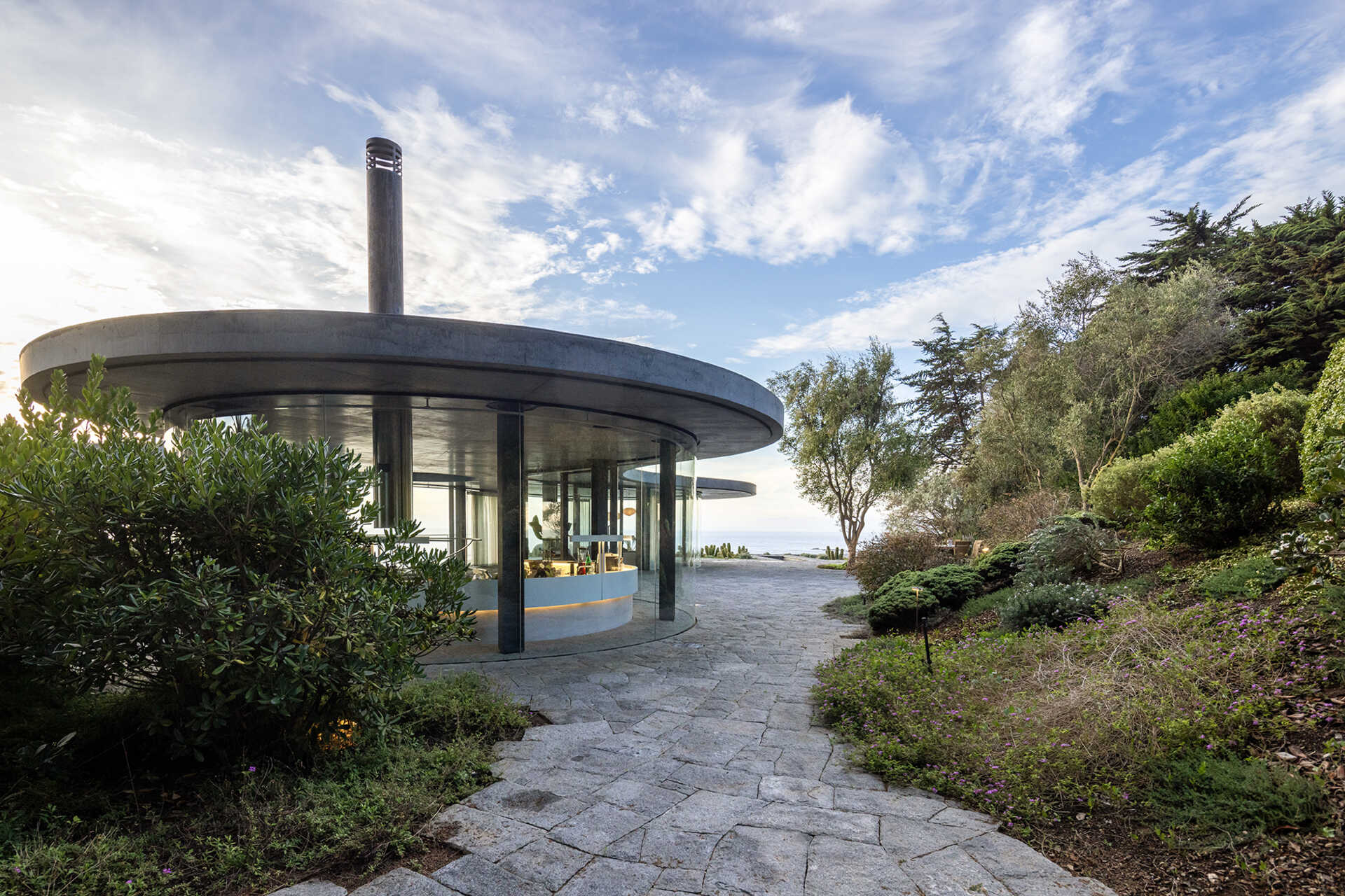 A concrete podium embedded into the landscape supports an amoeba-shaped glass pavilion, using stone, wood, and plank-form concrete throughout.