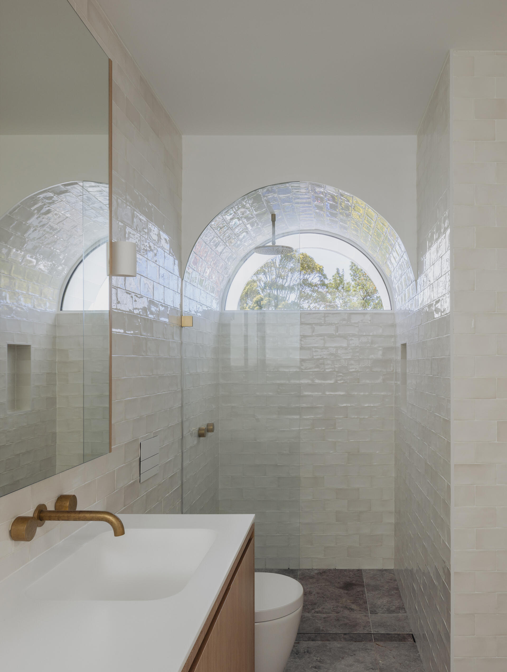In the bathroom, an arched window crowns the shower, drawing daylight deep into the space and framing soft views of greenery beyond. Glossy pale tiles wrap the walls, while a timber vanity and warm brass tapware introduce texture and warmth, balancing the crisp surfaces.