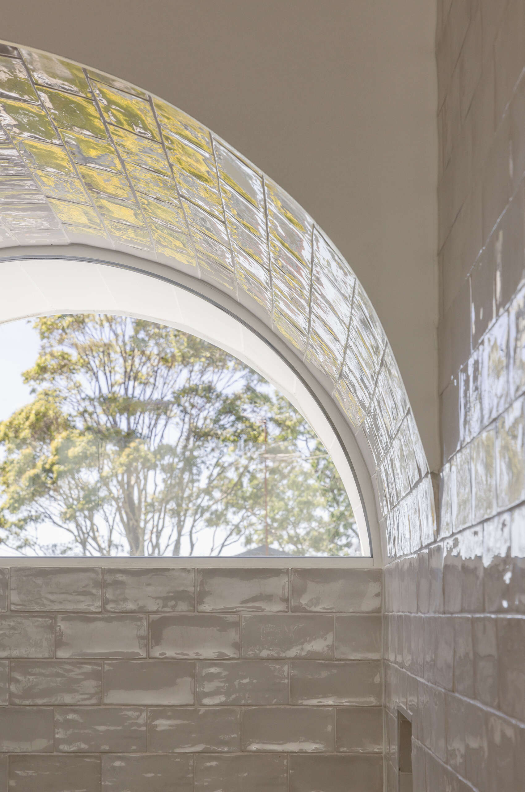 In the bathroom, an arched window crowns the shower, drawing daylight deep into the space and framing soft views of greenery beyond. Glossy pale tiles wrap the walls, while a timber vanity and warm brass tapware introduce texture and warmth, balancing the crisp surfaces.