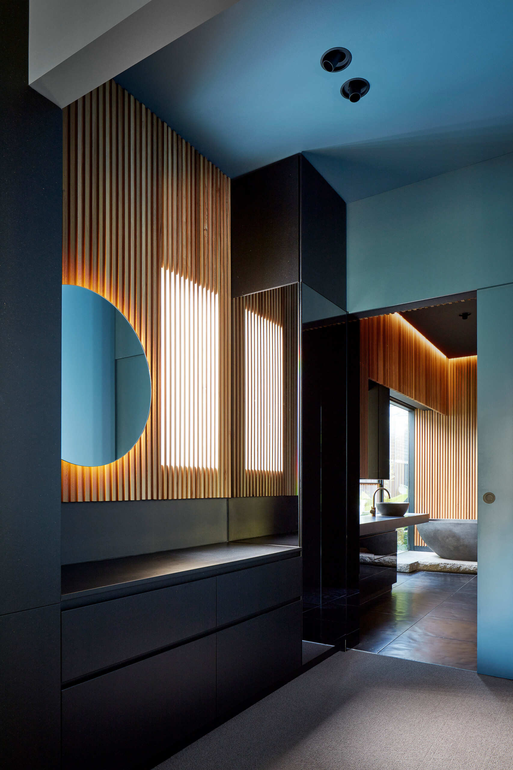 In the bathroom, concealed lighting creates a soft glow around the ceiling edges. Timber slats form a backdrop to black elements including the bathtub, floor tiles and window frame.