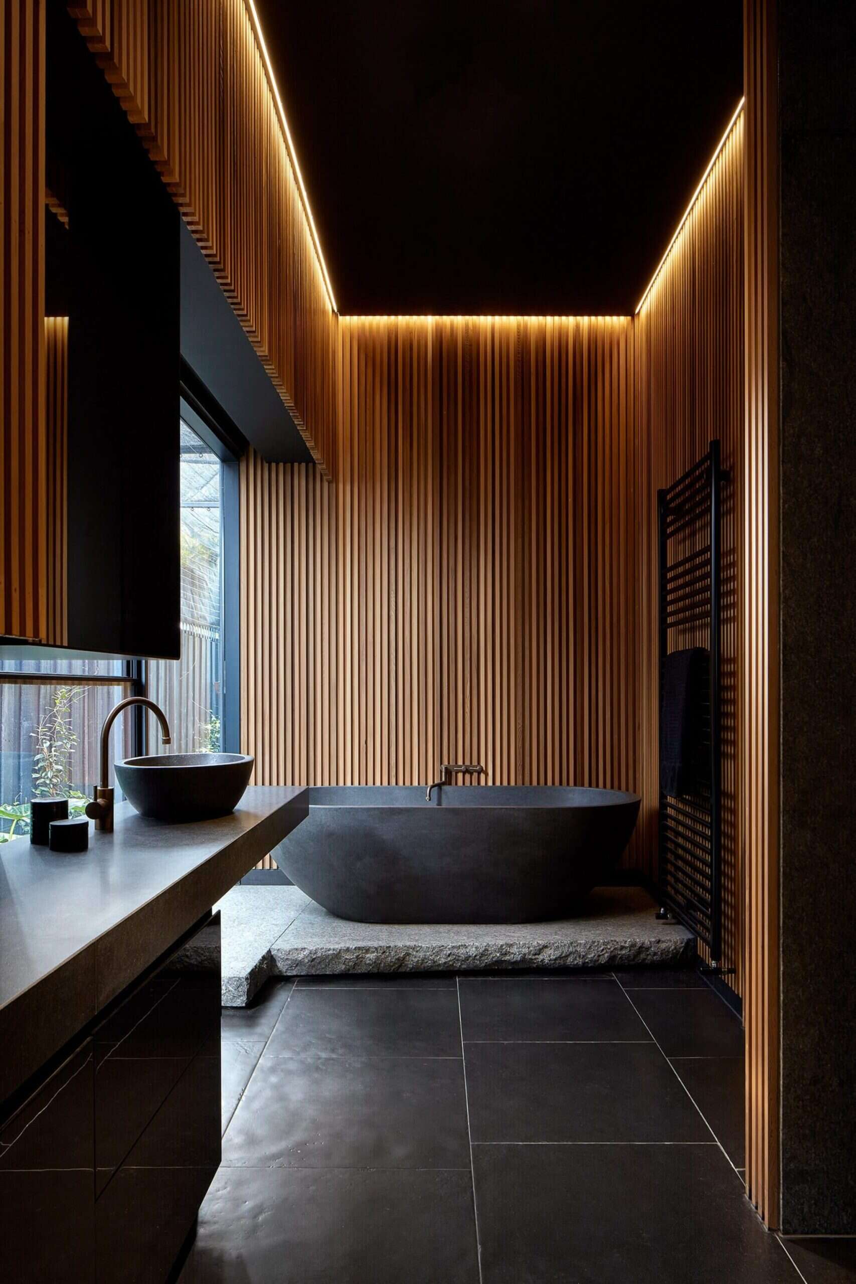 In the bathroom, concealed lighting creates a soft glow around the ceiling edges. Timber slats form a backdrop to black elements including the bathtub, floor tiles and window frame.