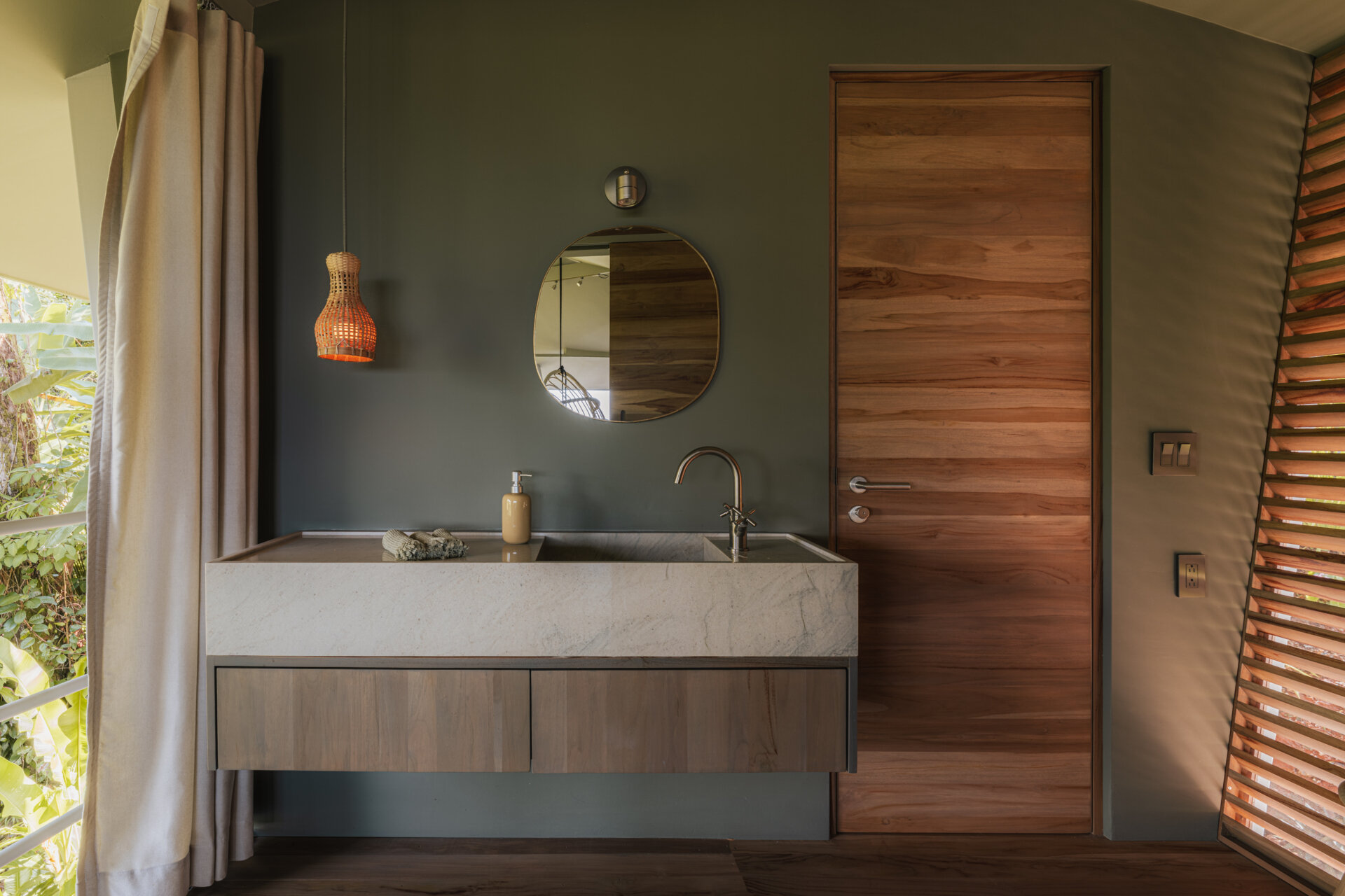The bathroom is pared back and calm, centered around a floating timber vanity topped with stone. Light filters through slatted timber and soft curtains, creating privacy while keeping the space open, tactile, and quietly connected to its surroundings.