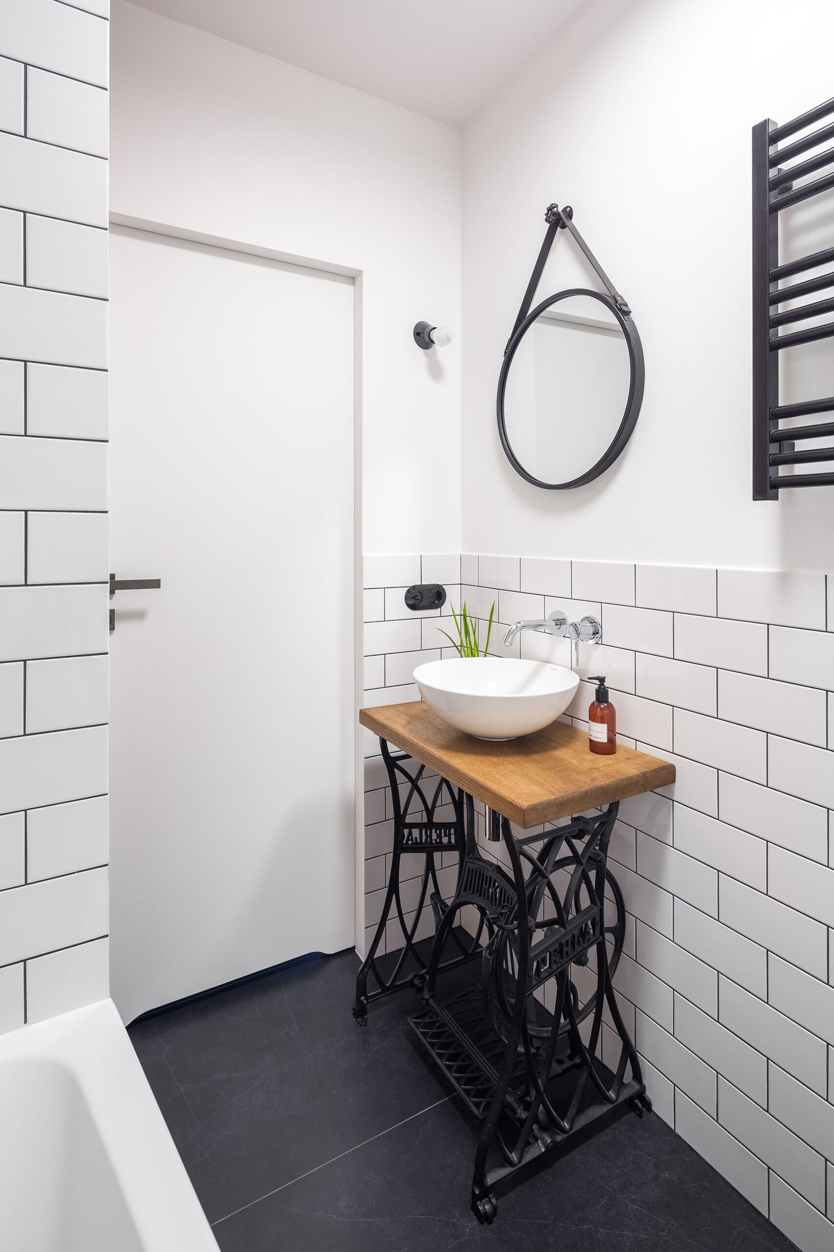 A sewing machine base vanity adds character to this small bathroom with black tiles and white subway walls.