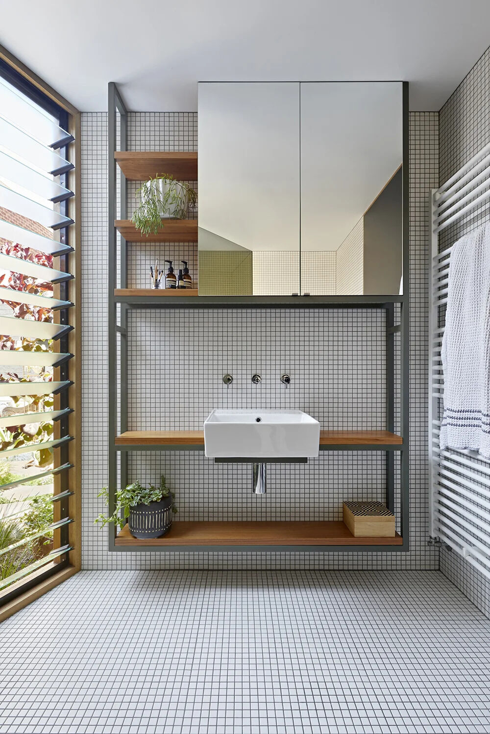 Timber shelving, an integrated mirror, and louvre windows create a bathroom that feels bright, functional, and serene.