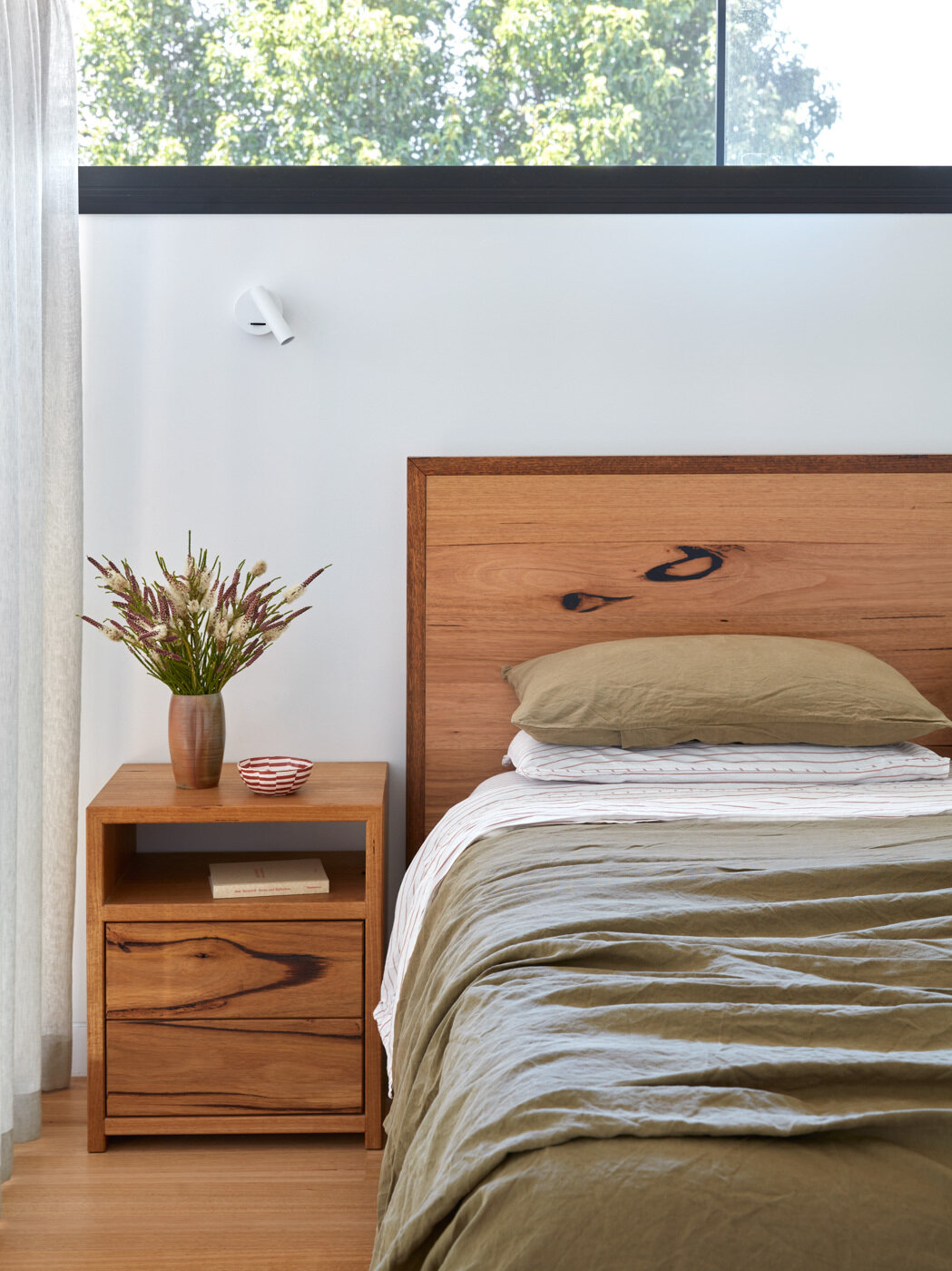 Este dormitorio equilibra la ligereza con la calma. Los cálidos muebles de madera y la suave ropa de cama de color verde oliva se apoyan en las paredes blancas, lo que permite que la vegetación exterior ocupe un lugar central. Las ventanas con marcos negros y los techos abovedados aportan una estructura tranquila, mientras que las cortinas transparentes y los biombos filtran la luz del sol y brindan privacidad.