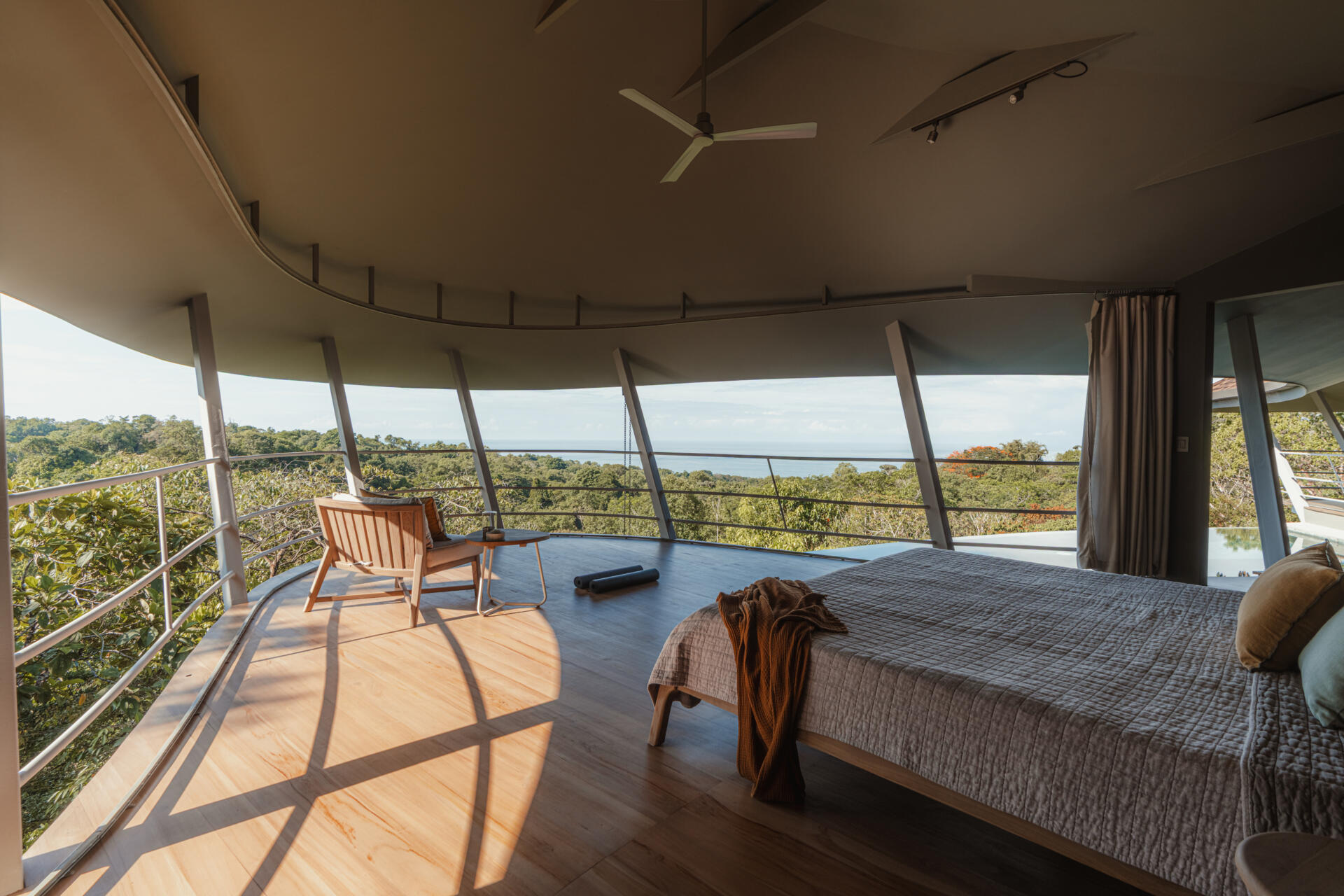 A curved open air bedroom where the sweeping roofline and angled supports frame uninterrupted views of forest and ocean. The low timber bed and minimal furnishings keep the focus on light, air, and landscape.