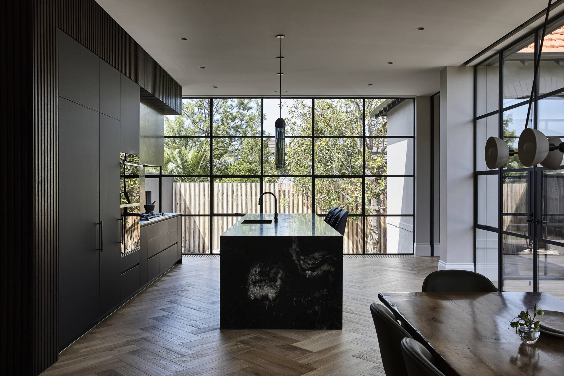 A sleek kitchen with matte black cabinetry, ribbed timber details, and dramatic black stone surfaces. Oversized grid windows and herringbone wood floors bring warmth and garden views inside.
