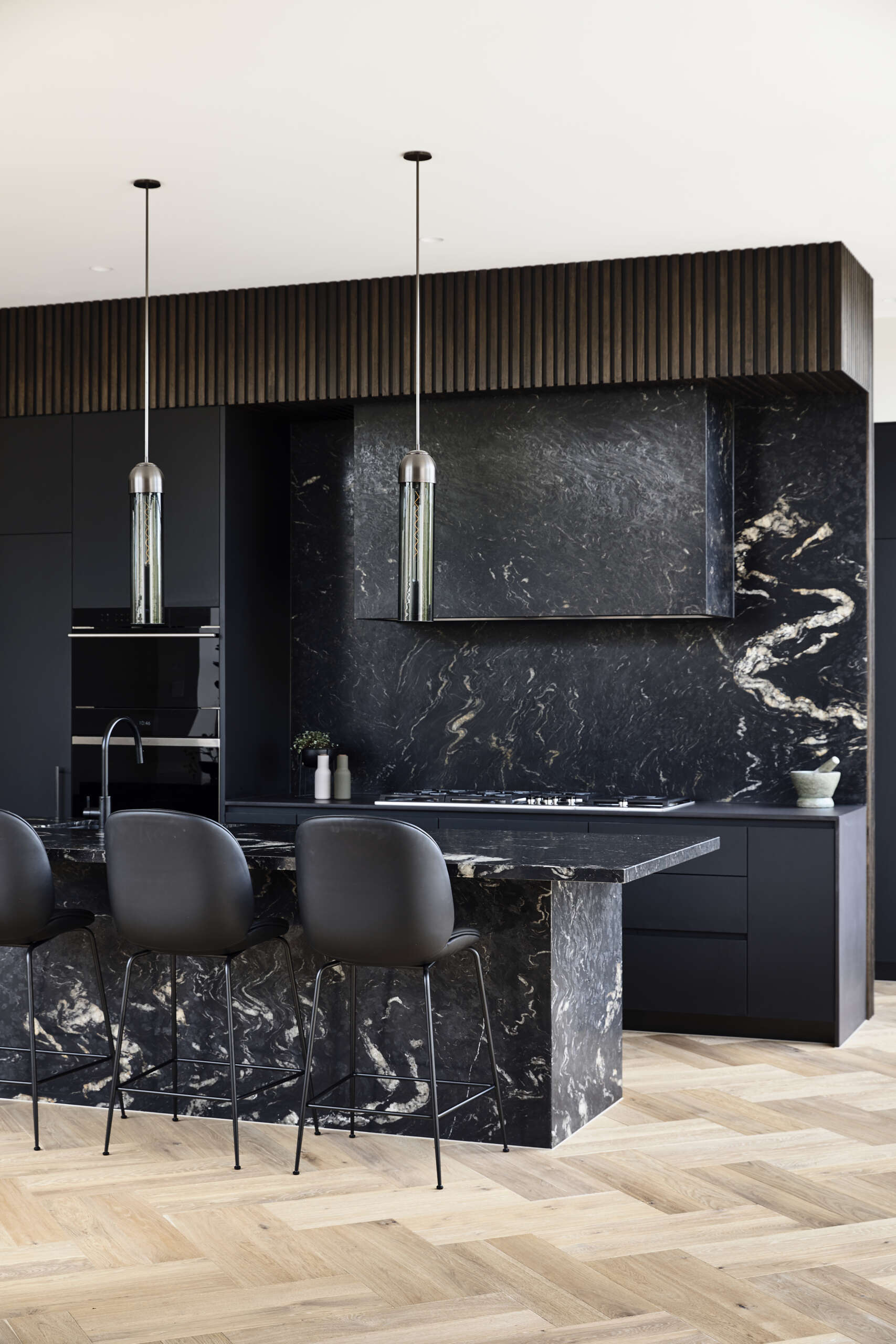 A sleek kitchen with matte black cabinetry, ribbed timber details, and dramatic black stone surfaces. Oversized grid windows and herringbone wood floors bring warmth and garden views inside.