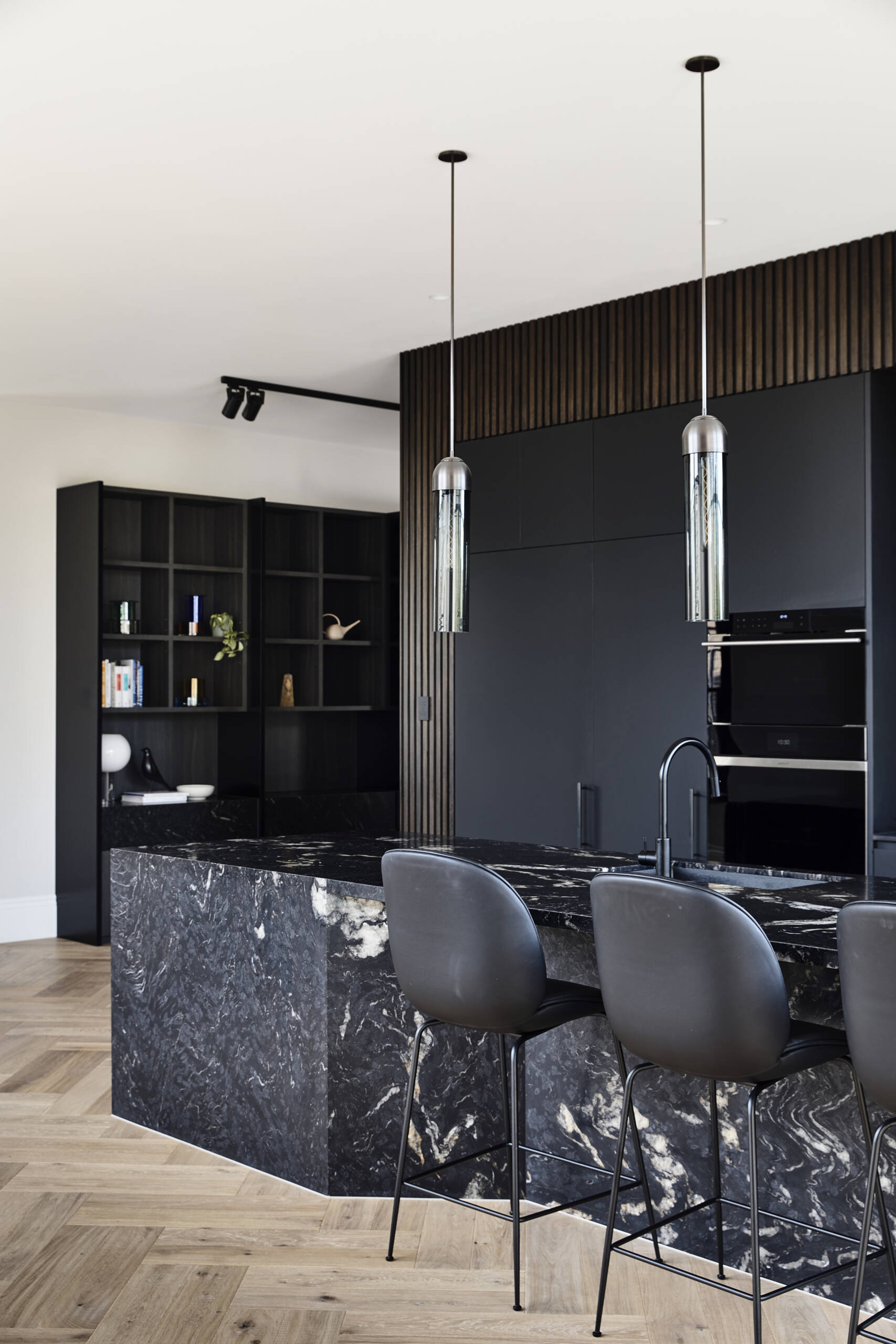 A sleek kitchen with matte black cabinetry, ribbed timber details, and dramatic black stone surfaces. Oversized grid windows and herringbone wood floors bring warmth and garden views inside.