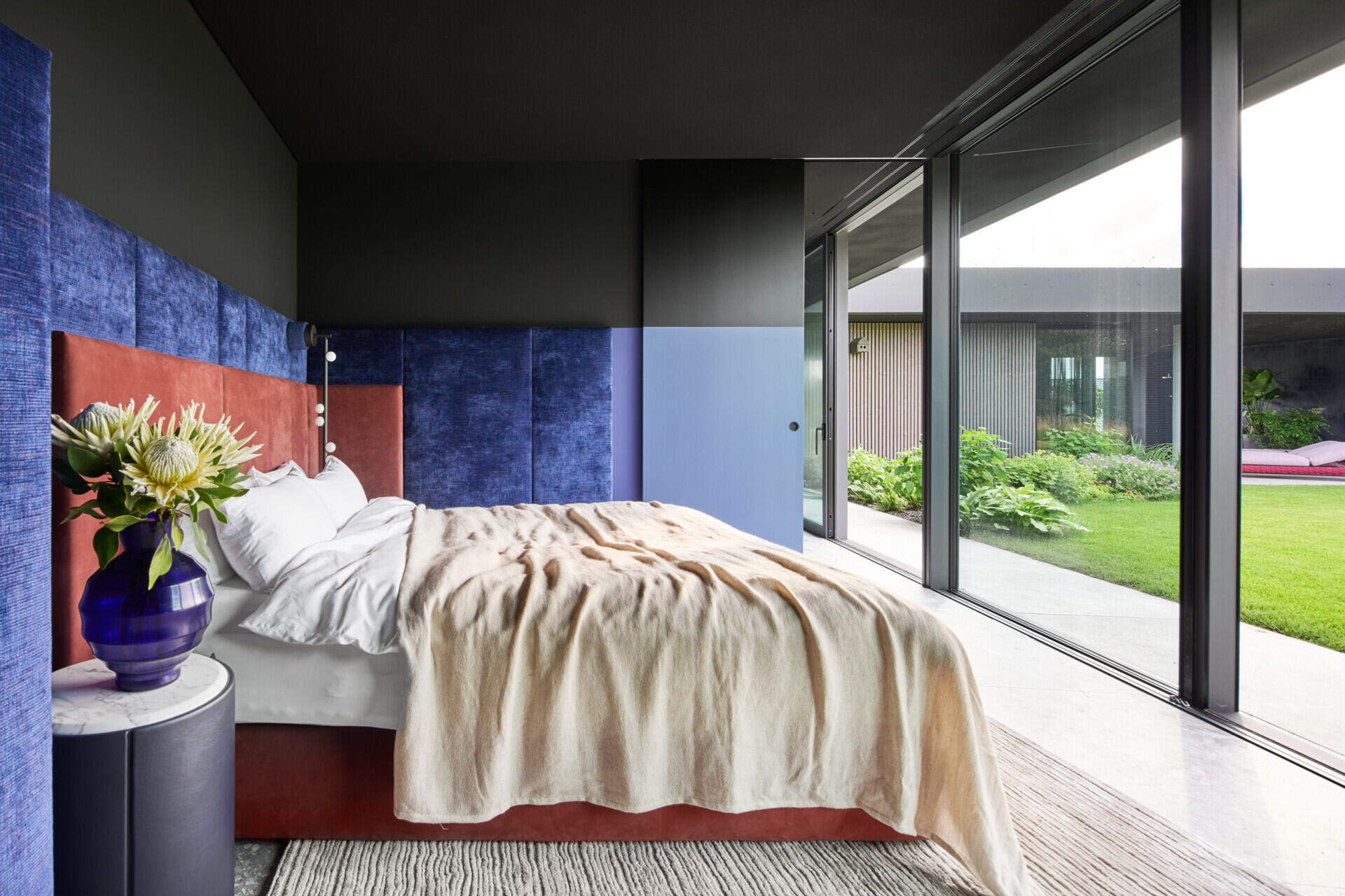 This bedroom and en-suite bathroom has a touch of drama, thanks to the velvety, ocean-blue wall panels. The rust-toned upholstered bed warms up the palette, while floor-to-ceiling glass opens straight to the garden. Nearby, the en-suite bathroom features a built-in bathtub with terrazzo walls and floors.