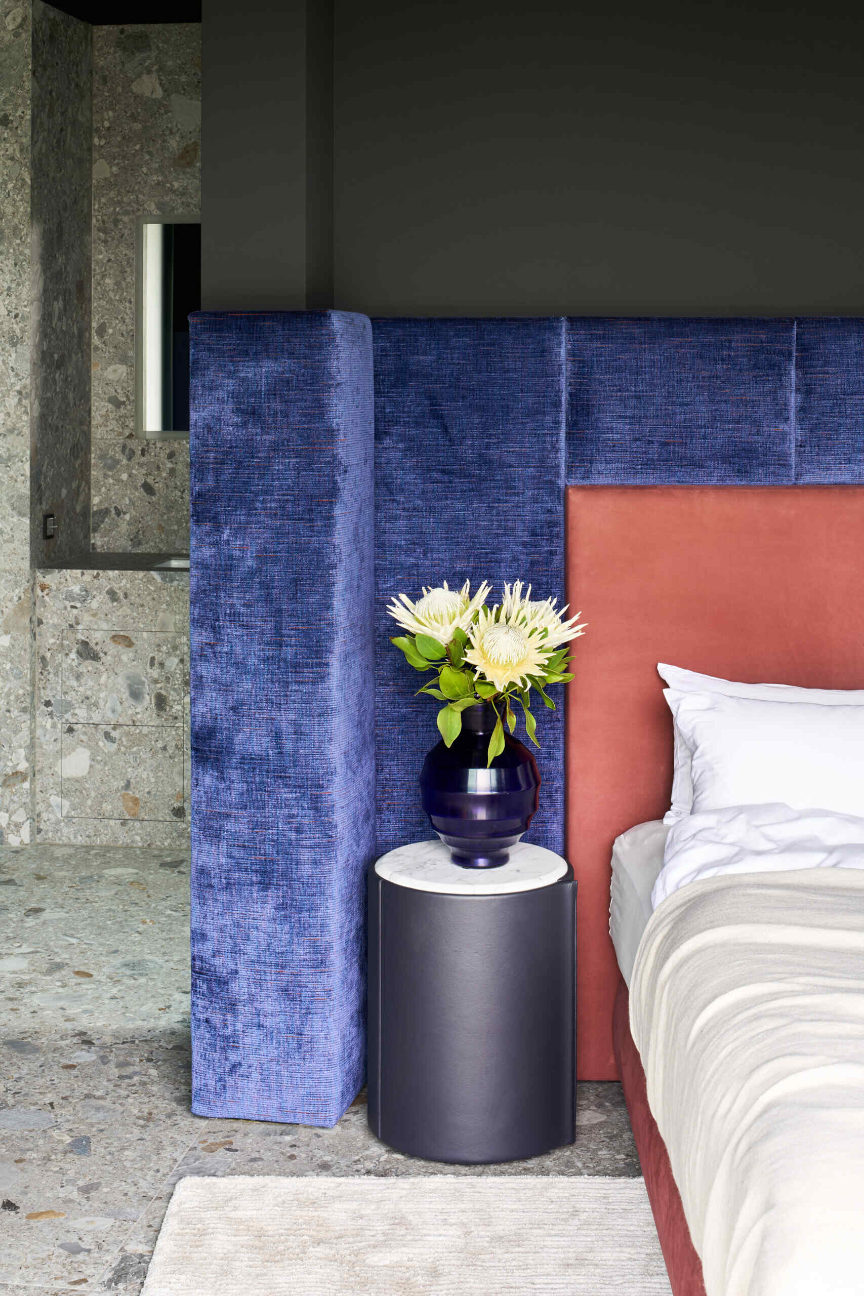 This bedroom and en-suite bathroom has a touch of drama, thanks to the velvety, ocean-blue wall panels. The rust-toned upholstered bed warms up the palette, while floor-to-ceiling glass opens straight to the garden. Nearby, the en-suite bathroom features a built-in bathtub with terrazzo walls and floors.