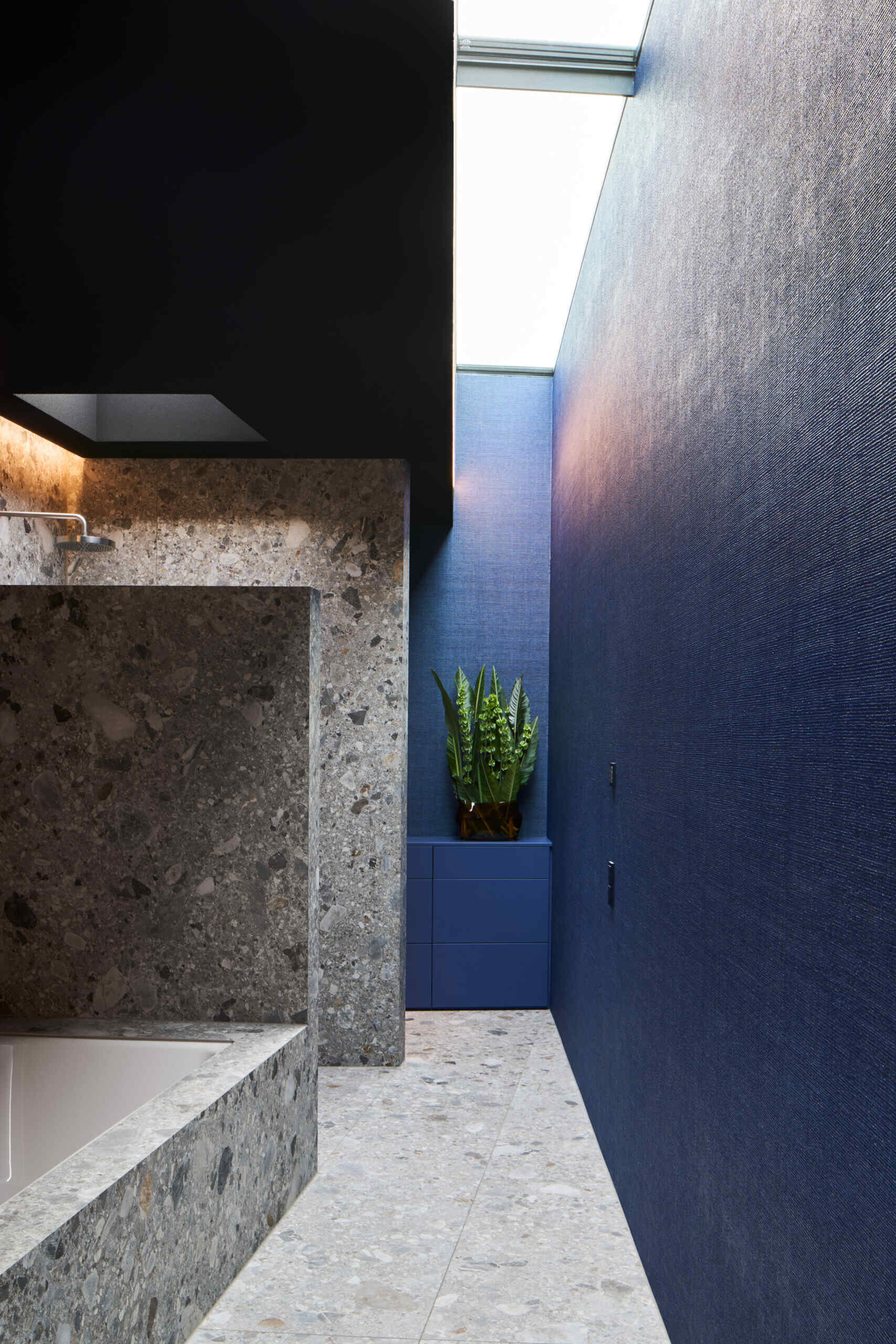 This bedroom and en-suite bathroom has a touch of drama, thanks to the velvety, ocean-blue wall panels. The rust-toned upholstered bed warms up the palette, while floor-to-ceiling glass opens straight to the garden. Nearby, the en-suite bathroom features a built-in bathtub with terrazzo walls and floors.