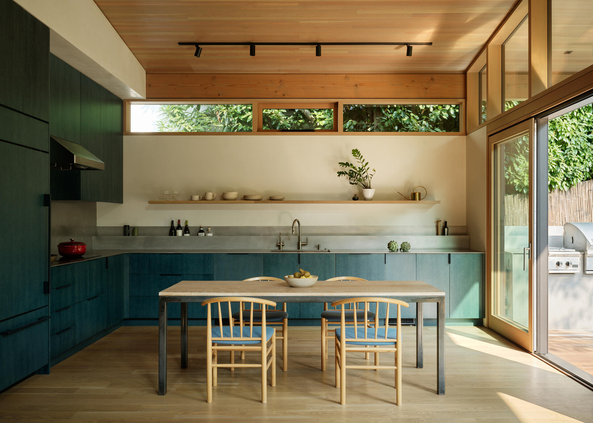 Deep petrol-blue cabinets define the kitchen, while white oak, plaster walls, a metal countertop, and natural wood floors make the space both practical and visually engaging, keeping it connected to the overall home aesthetic.