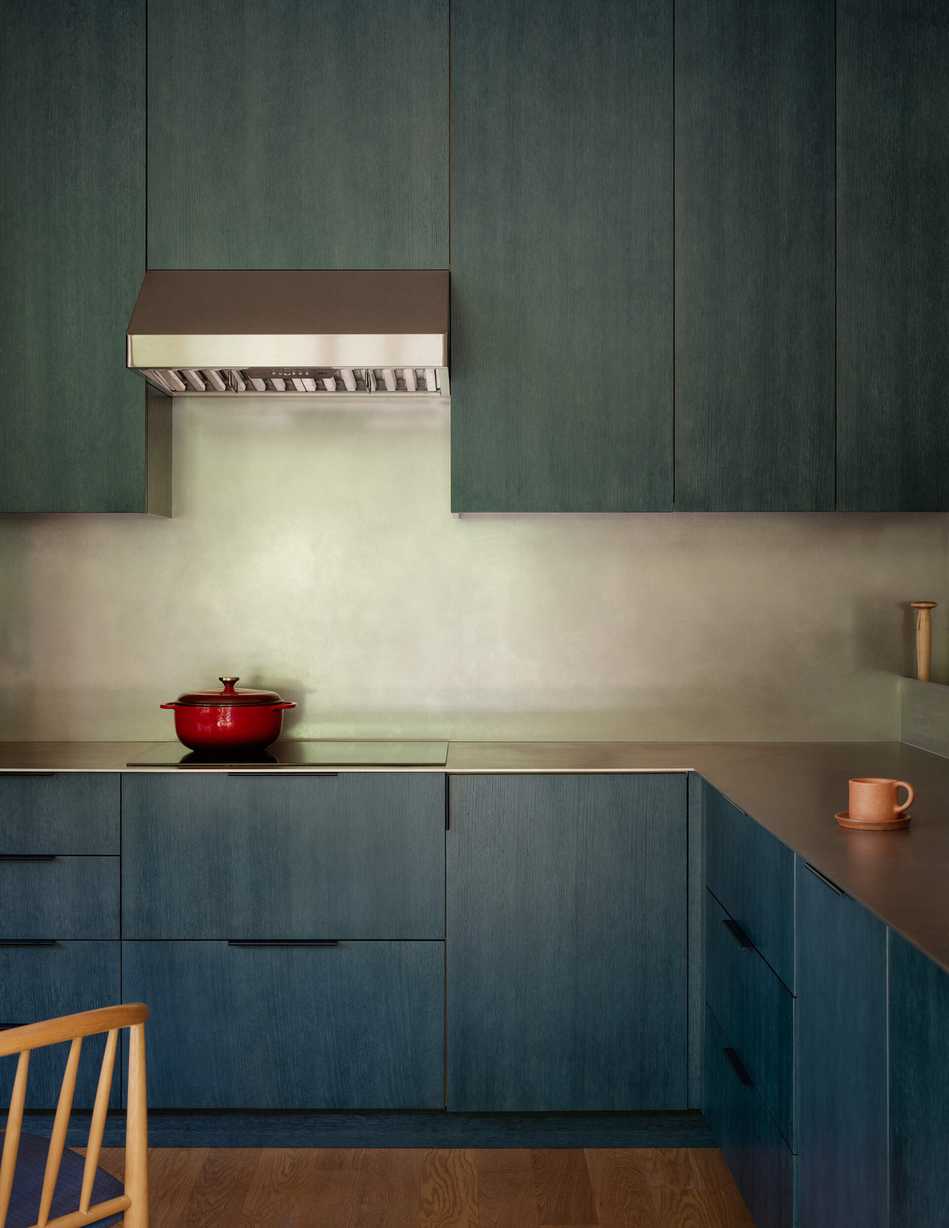 Deep petrol-blue cabinets define the kitchen, while white oak, plaster walls, a metal countertop, and natural wood floors make the space both practical and visually engaging, keeping it connected to the overall home aesthetic.