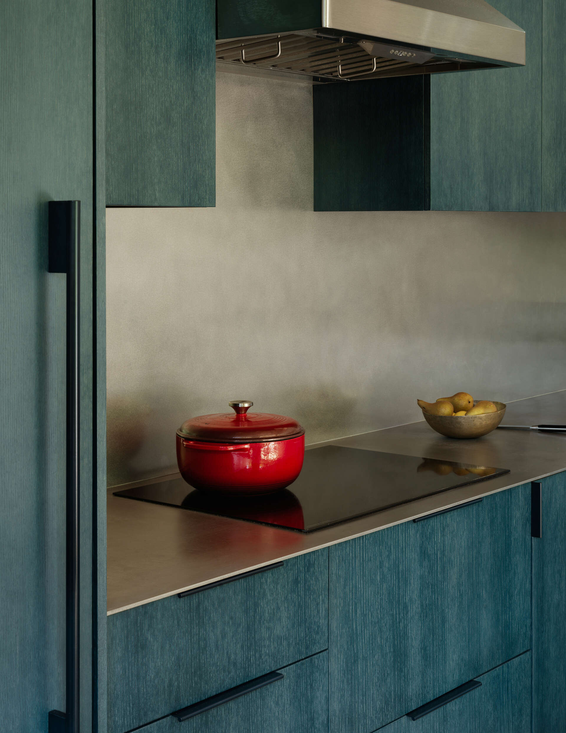 Deep petrol-blue cabinets define the kitchen, while white oak, plaster walls, a metal countertop, and natural wood floors make the space both practical and visually engaging, keeping it connected to the overall home aesthetic.