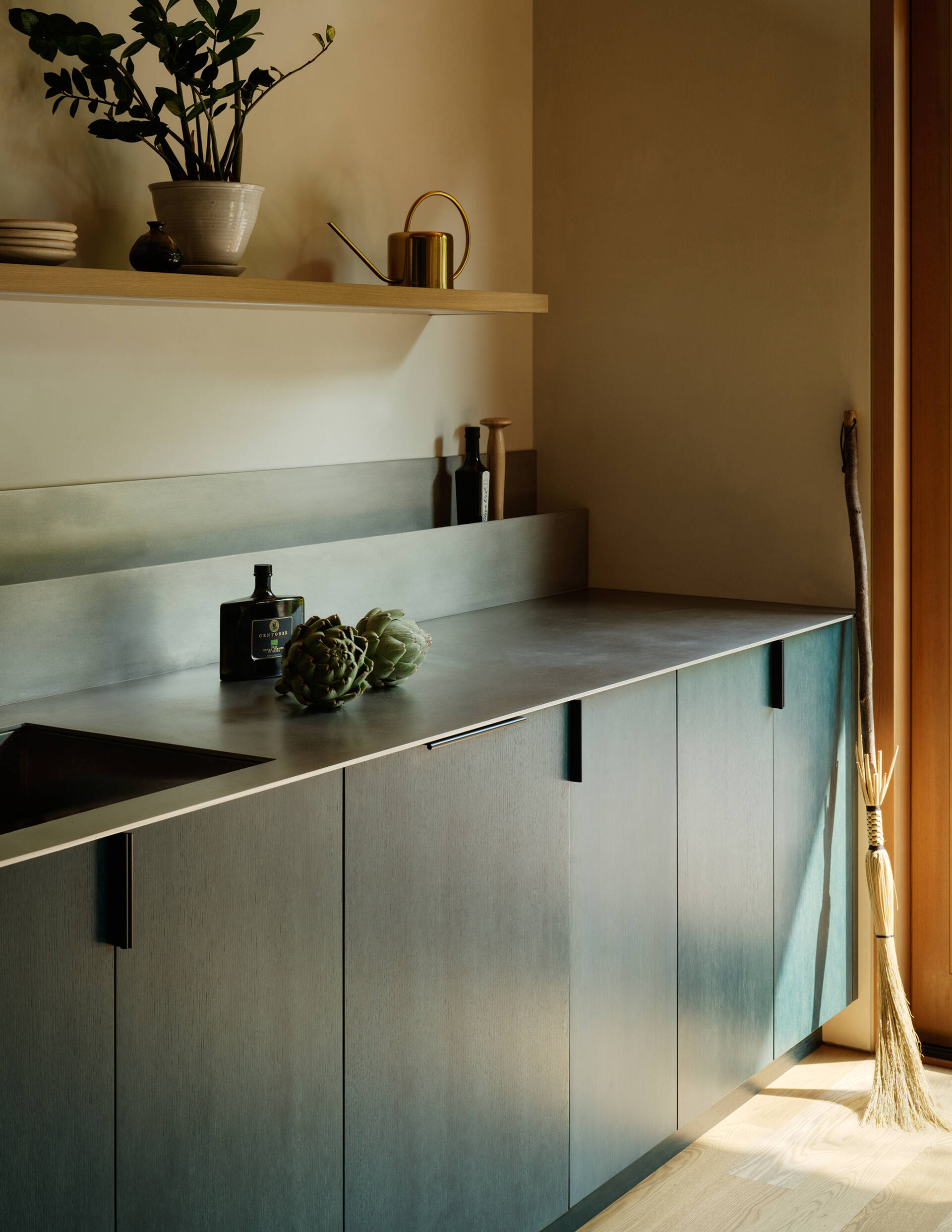 Deep petrol-blue cabinets define the kitchen, while white oak, plaster walls, a metal countertop, and natural wood floors make the space both practical and visually engaging, keeping it connected to the overall home aesthetic.