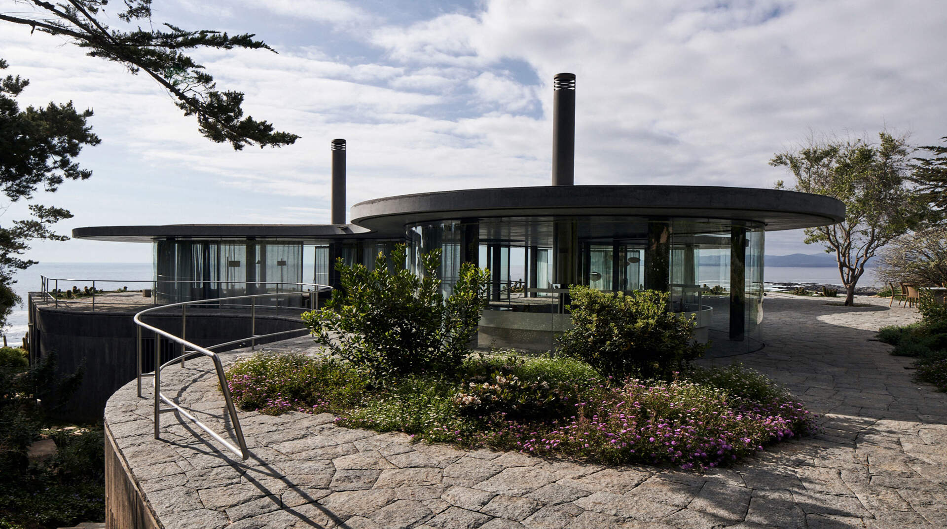 A concrete podium embedded into the landscape supports an amoeba-shaped glass pavilion, using stone, wood, and plank-form concrete throughout.