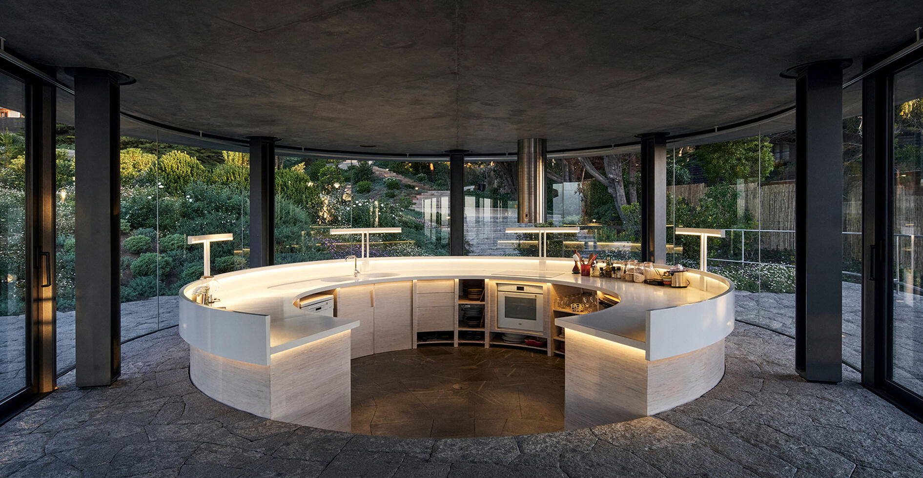 An open curved kitchen within the glass pavilion where structure fades and the coastal view becomes the focus.
