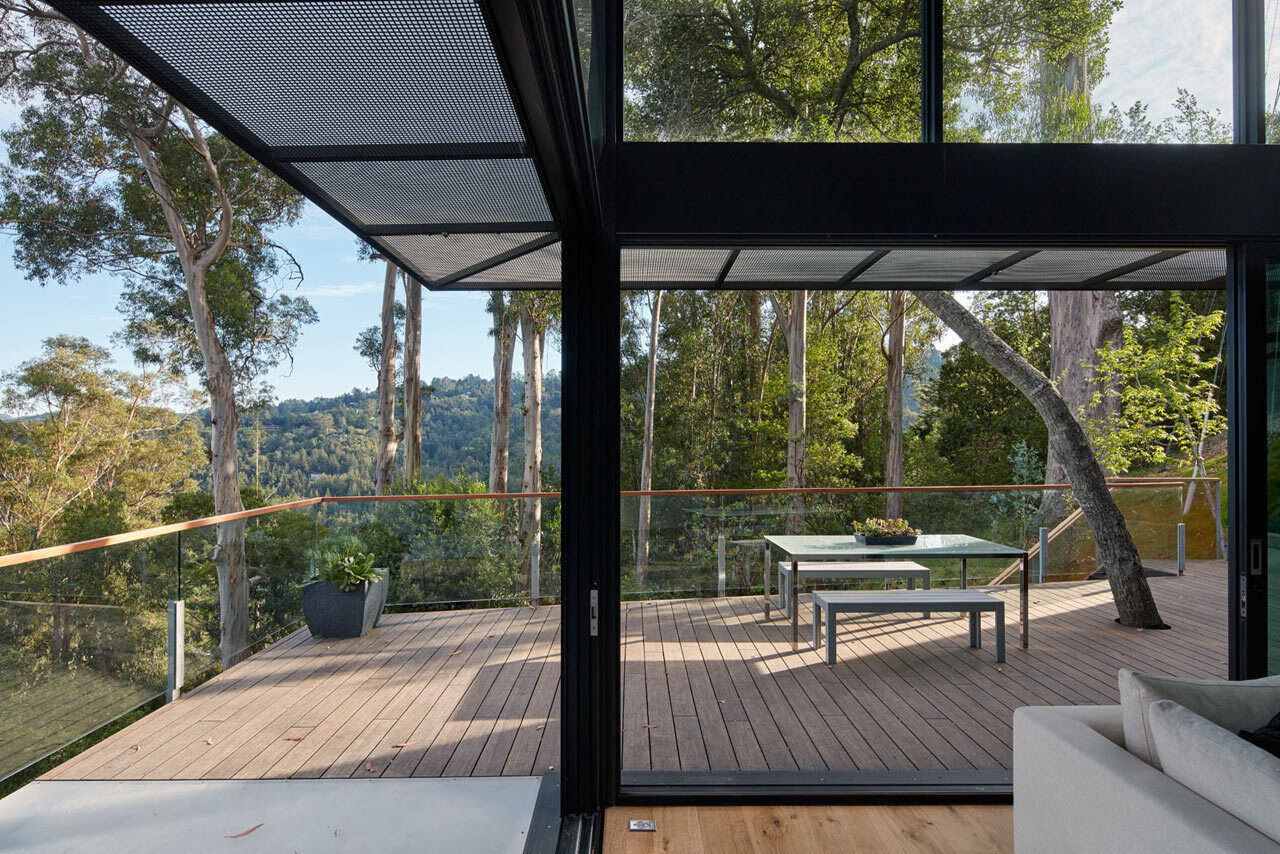 Las puertas corredizas de vidrio abren la sala de estar a una terraza con barandillas de vidrio que ofrecen vistas despejadas de los árboles y las laderas circundantes.