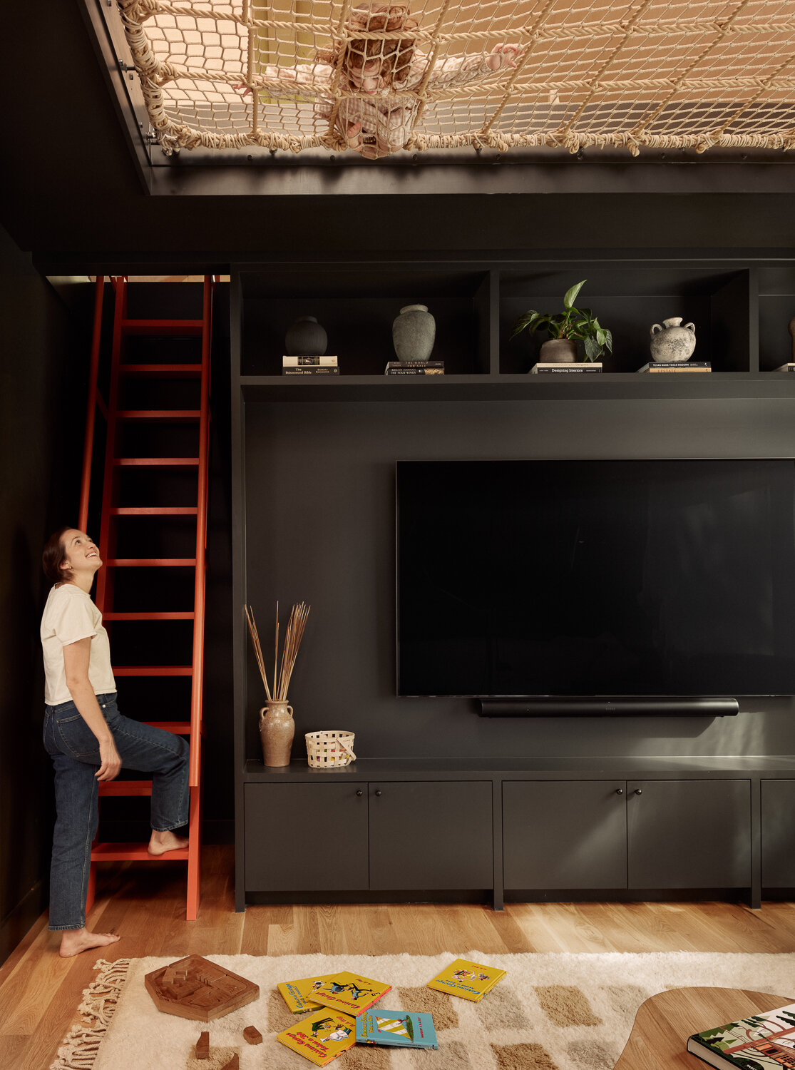 Back inside, and the family room is designed for comfort and everyday use. A matte black built-in wall anchors the space and creates a relaxed, movie-night atmosphere, with open cubbies styled in pottery and greenery. A bright red ladder adds a playful note and leads up to a rope net loft and play area, turning vertical space into an extension of the room.