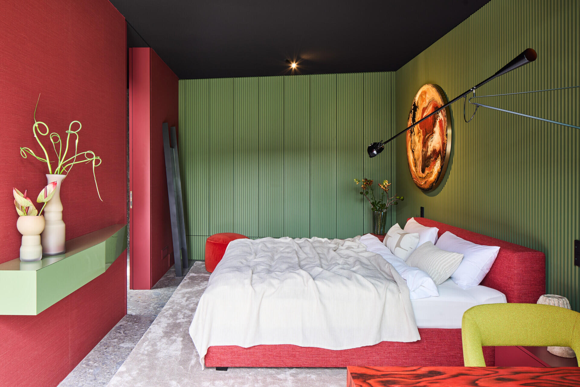 Bold color blocking gives this bedroom instant energy, with red and ribbed olive-green walls wrapping the space. A plush red upholstered bed anchors the room, softened by cloud-white bedding, while the round amber artwork and simple vases bring personality without clutter.