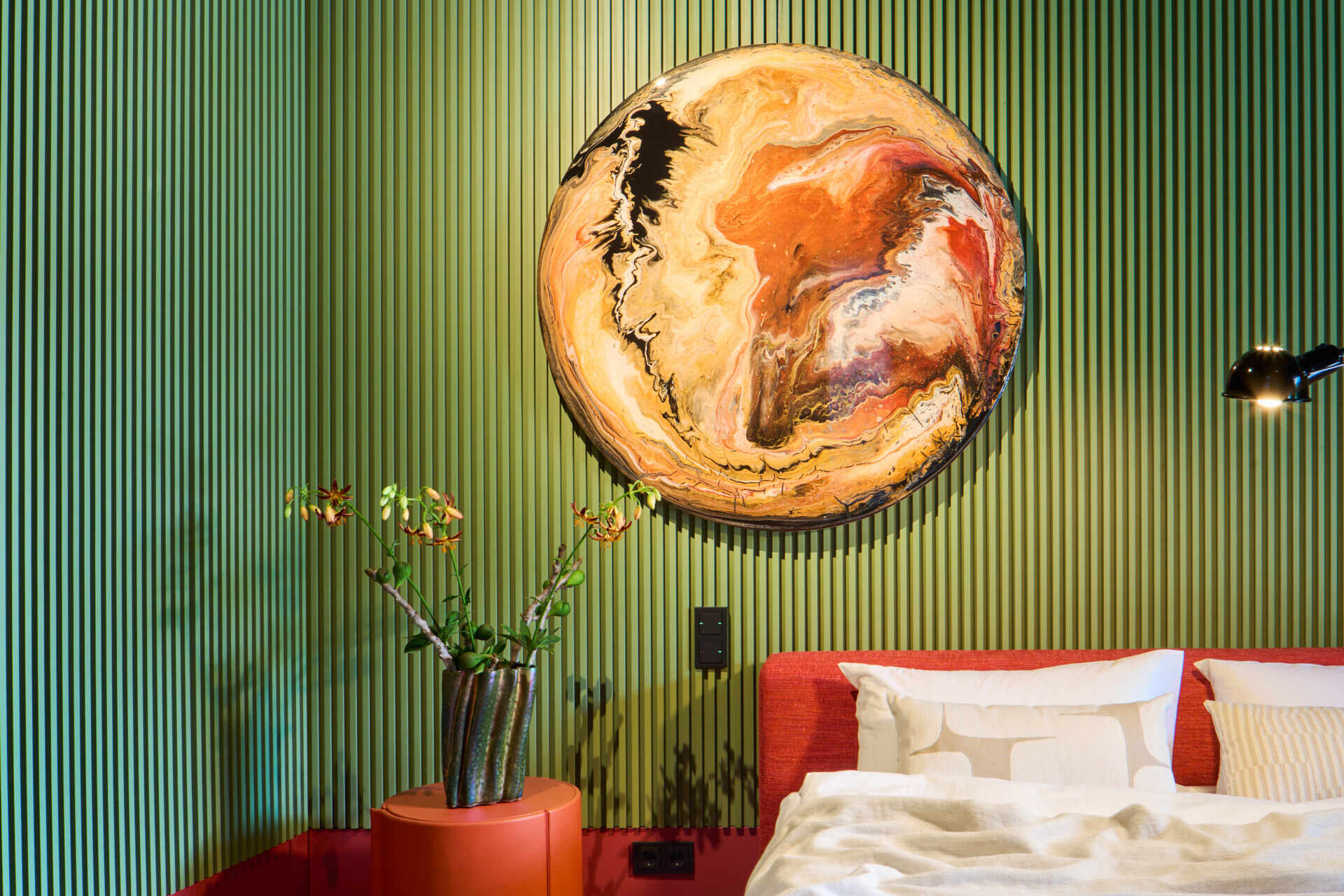 Bold color blocking gives this bedroom instant energy, with red and ribbed olive-green walls wrapping the space. A plush red upholstered bed anchors the room, softened by cloud-white bedding, while the round amber artwork and simple vases bring personality without clutter.