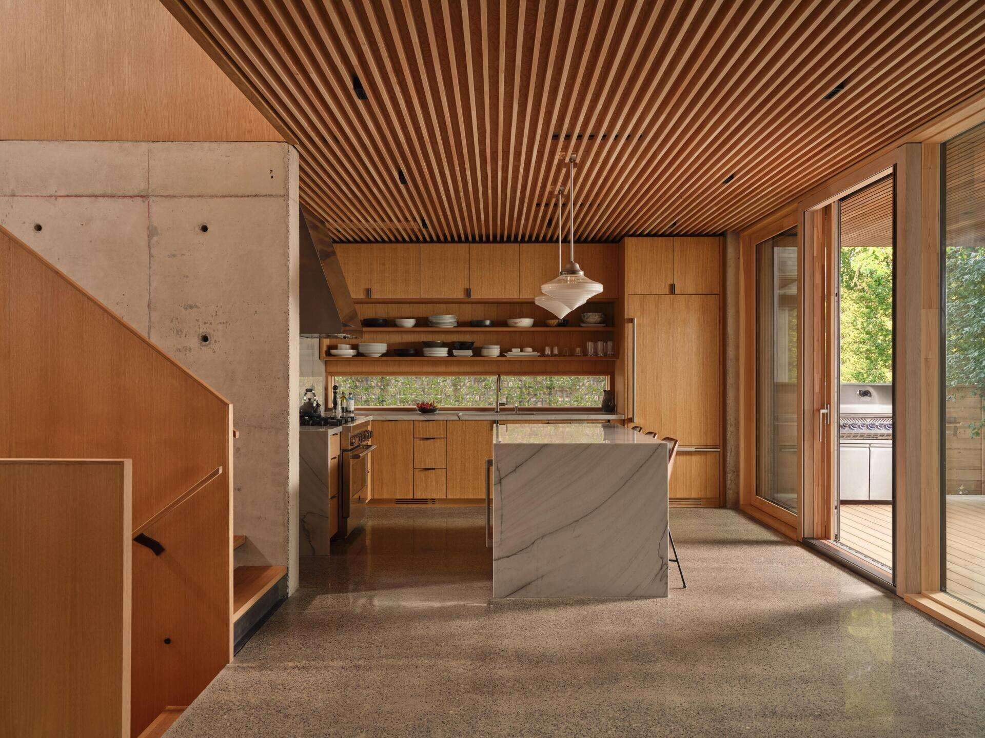 The kitchen sits within the concrete framework, unified with the living room by a Douglas Fir ceiling. Oak millwork reinforces the continuity between spaces while remaining clearly inserted into the structural shell. From here, views extend south toward Lake Ontario, with light shaping both movement and pause within the room.