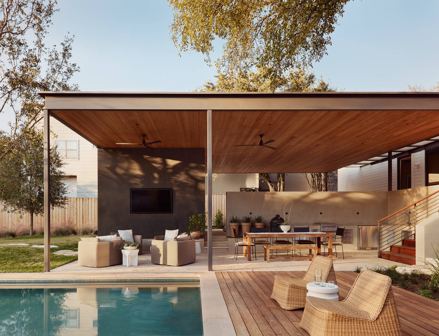 Outside, the pool and deck area extends the home’s living space outdoors. A wide, flat roof provides shade over both lounging and dining areas, making the space comfortable throughout the day. Warm wood ceilings and decking soften the modern structure, while a charcoal feature wall with a mounted television adds a cozy focal point for evenings. Woven chaise loungers sit close to the pool, and a built-in grill with a long dining table makes hosting feel effortless.