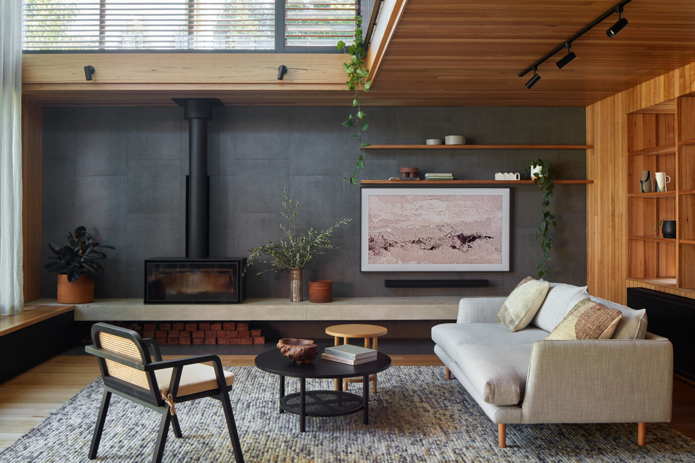 La sala de estar cuenta con cálidas paredes y techos de madera rodeados por una profunda chimenea de carbón, lo que crea un telón de fondo cómodo y moderno para asientos suaves y neutros. La luz del sol se cuela a través del alto vidrio con marco negro y las cortinas transparentes suavizan la luz, manteniendo la vista brillante y suave.