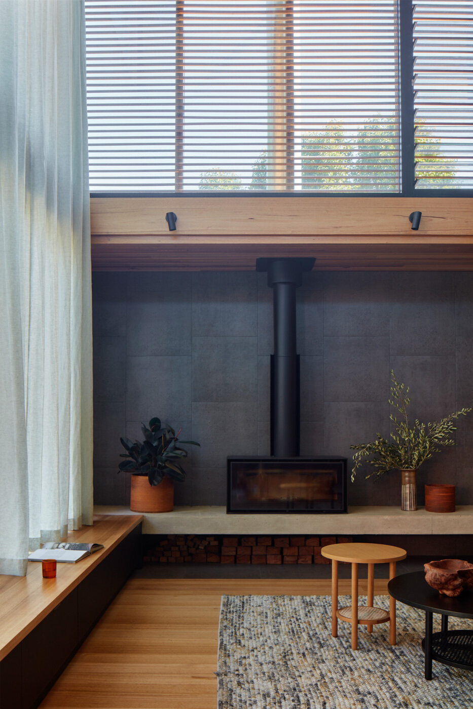 La sala de estar cuenta con cálidas paredes y techos de madera rodeados por una profunda chimenea de carbón, lo que crea un telón de fondo cómodo y moderno para asientos suaves y neutros. La luz del sol se cuela a través del alto vidrio con marco negro y las cortinas transparentes suavizan la luz, manteniendo la vista brillante y suave.