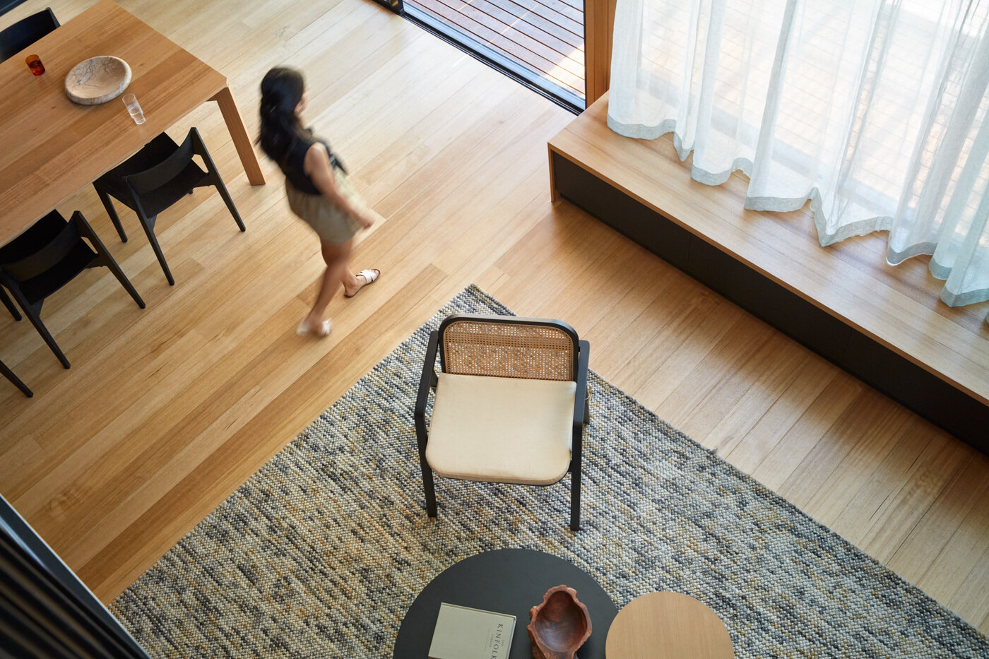 La sala de estar cuenta con cálidas paredes y techos de madera rodeados por una profunda chimenea de carbón, lo que crea un telón de fondo cómodo y moderno para asientos suaves y neutros. La luz del sol se cuela a través del alto vidrio con marco negro y las cortinas transparentes suavizan la luz, manteniendo la vista brillante y suave.
