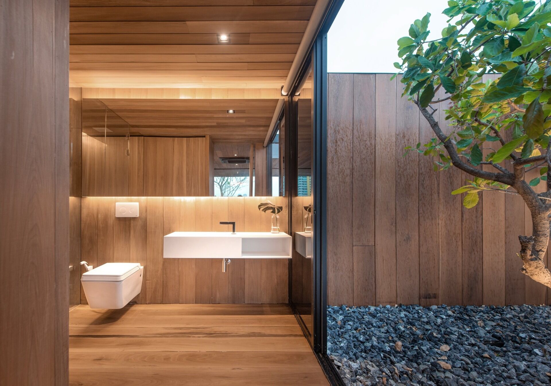 White accents, a backlit mirror, and a full height window bring light into the wood lined bathroom interior.