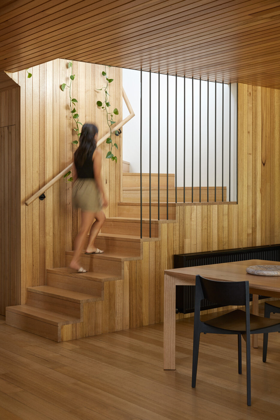 La madera en tonos miel envuelve las paredes, techos y escaleras, creando continuidad y profundidad. Las esbeltas barandillas negras crean un ritmo tranquilo, mientras que las plantas trepadoras suavizan los bordes y añaden vida. En el rellano, las paredes blancas reflejan la luz hacia el espacio.