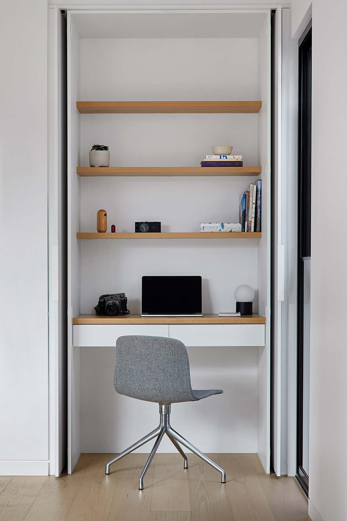 A compact home office is concealed within a closet, featuring warm wood shelving and a clean, minimal setup.