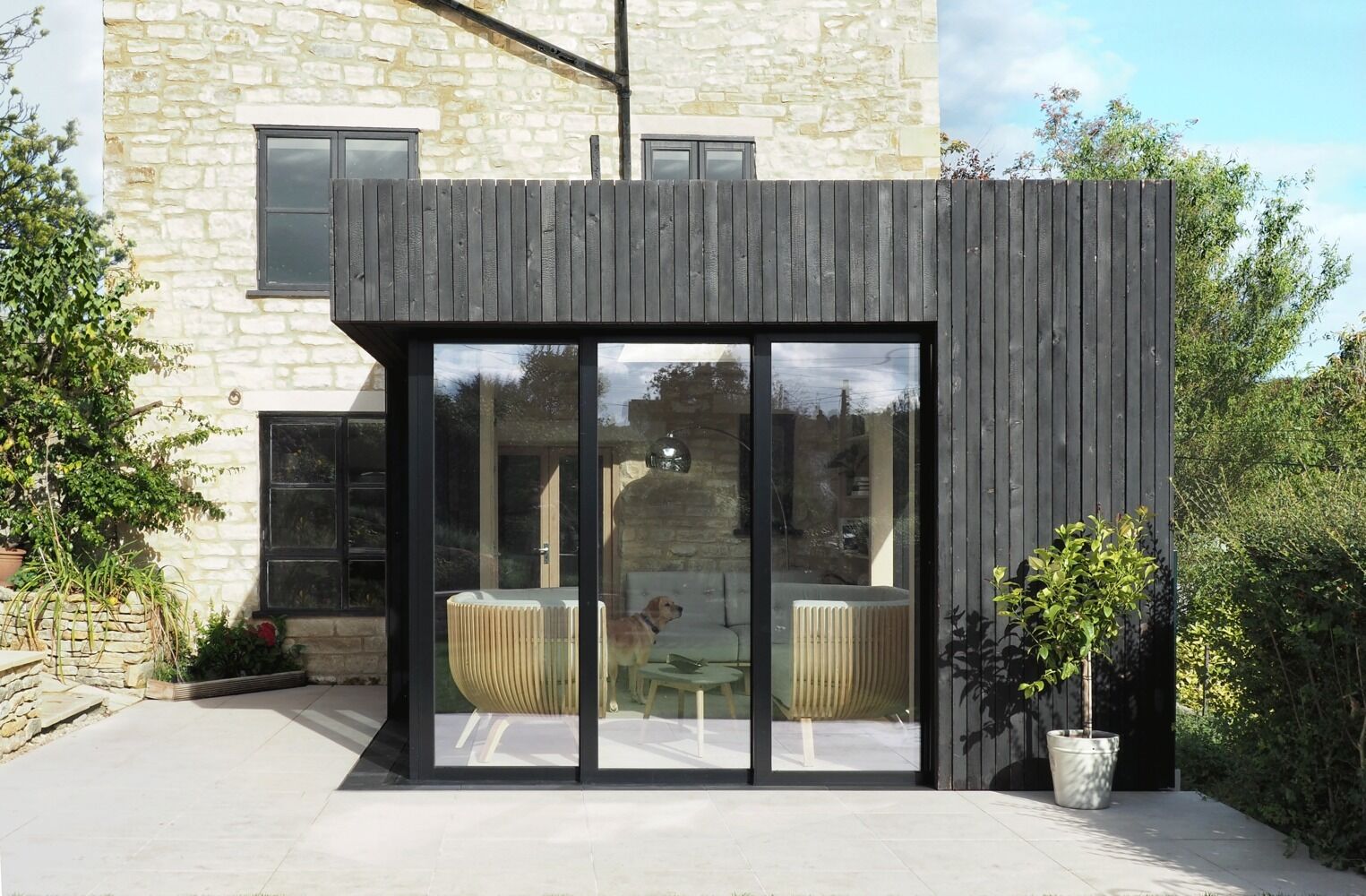 Una cabaña histórica de Cotswold combina un diseño moderno con vigas carbonizadas, marcos negros y paredes de vidrio que desaparecen y se abren directamente al jardín.