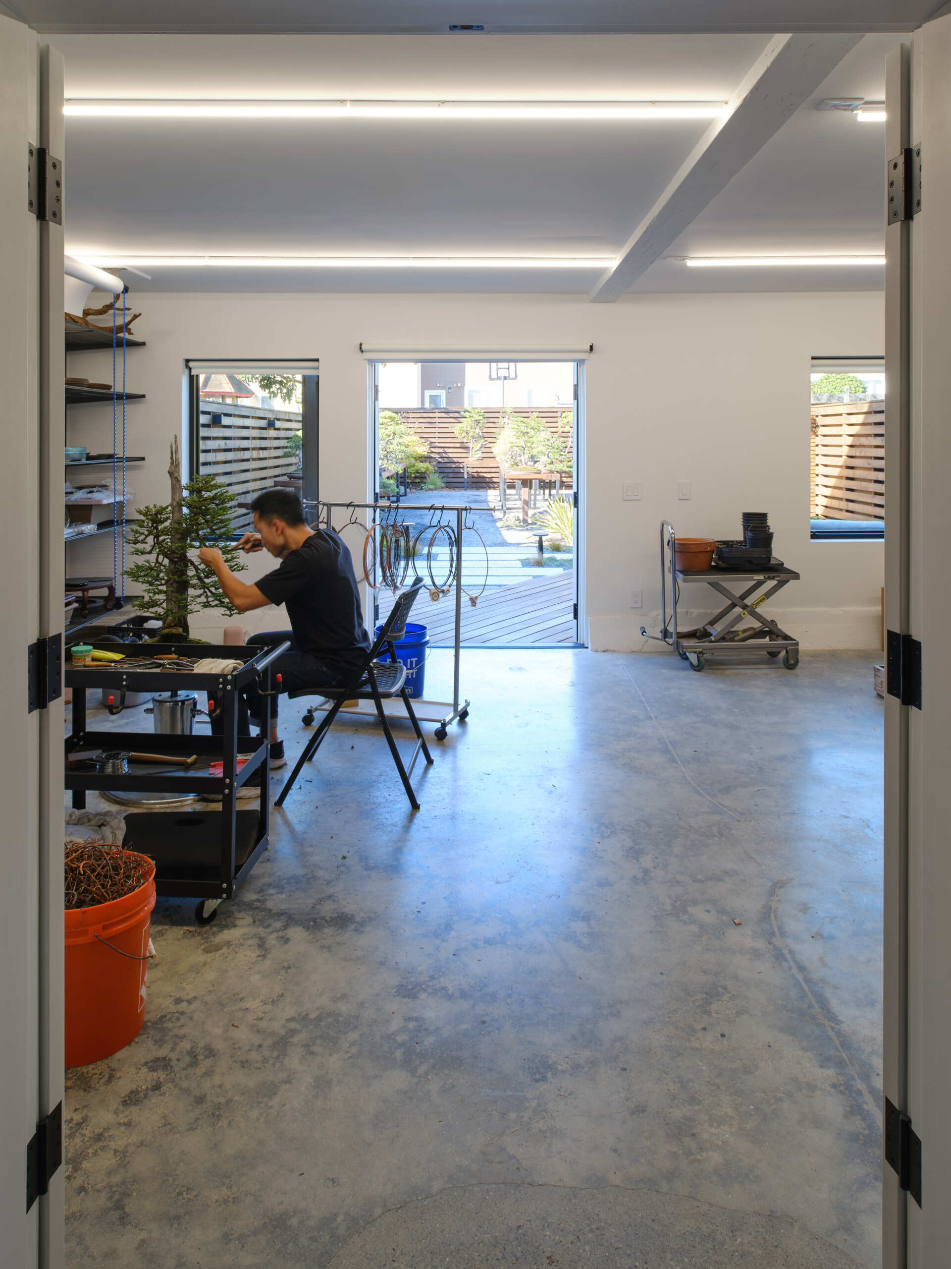 The renovation introduces a dedicated workshop environment with wide French doors opening from street to garden. Large carts can move bonsai trees directly from truck to backyard with ease, while still accommodating a single car garage.