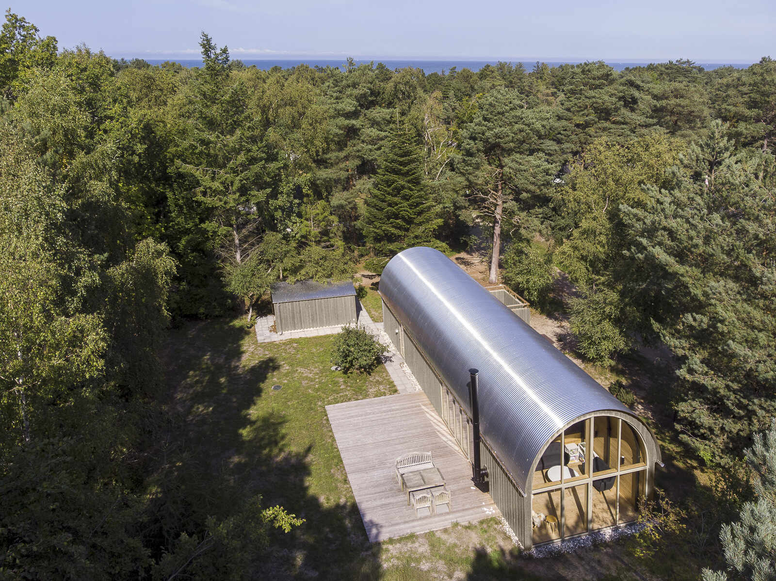 A tube-shaped Danish summer home set in the forest just 200 metres from the sea, featuring a curved corrugated steel roof and a bold minimalist form.