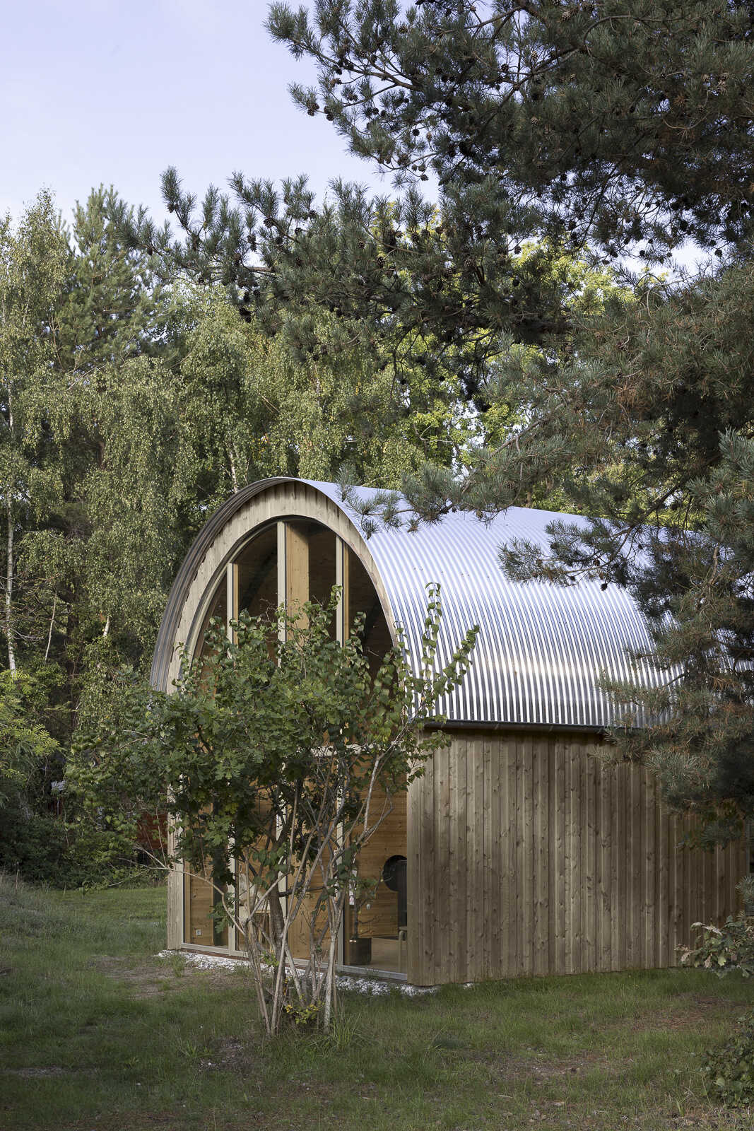 A tube-shaped Danish summer home set in the forest just 200 metres from the sea, featuring a curved corrugated steel roof and a bold minimalist form.