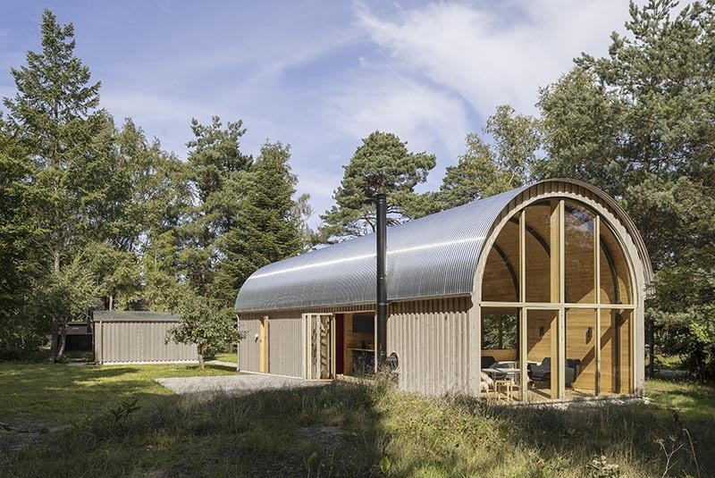 A tube-shaped Danish summer home set in the forest just 200 metres from the sea, featuring a curved corrugated steel roof and a bold minimalist form.
