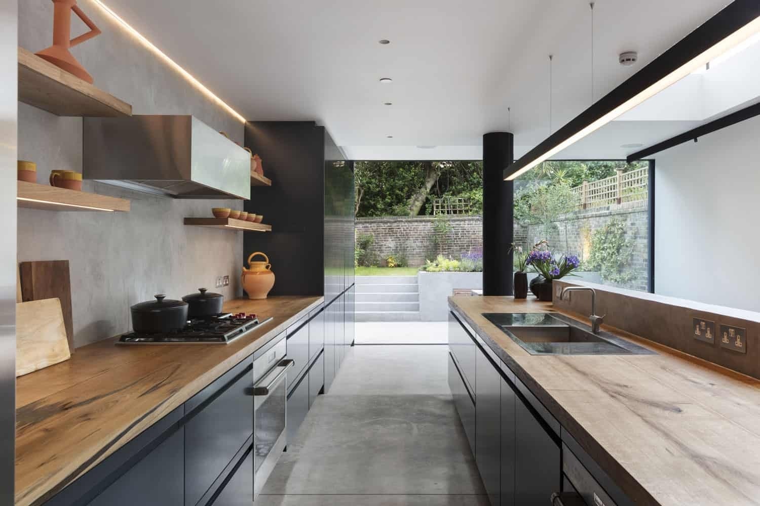 A modern kitchen with dark cabinets, concrete floors and oak countertops sits at the heart of this open plan glass extension designed for entertaining.